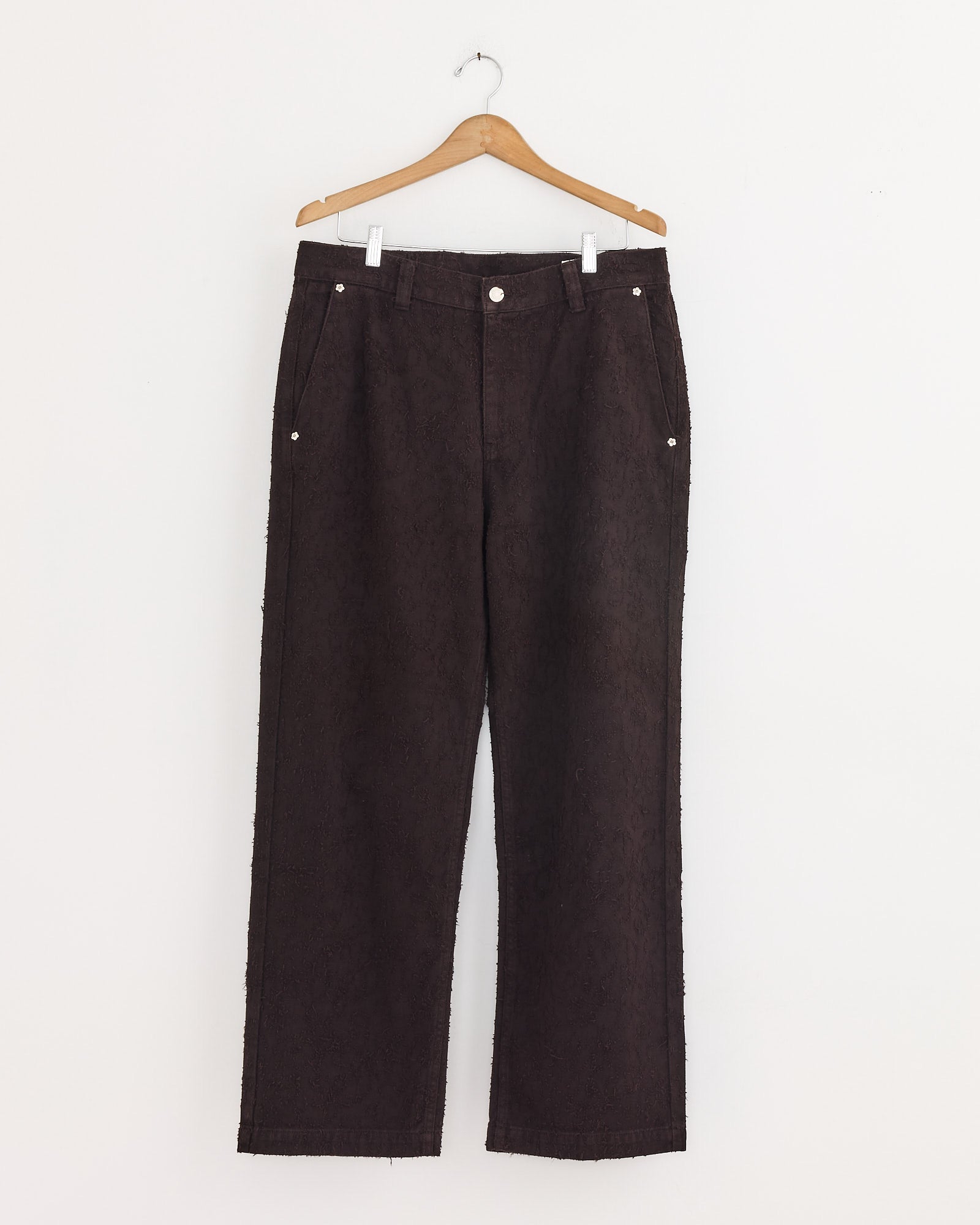 The mfpen Punch Out Jean in Dark Brown is a high-waisted, straight-leg style made from organic cotton denim with a vintage finish, silver rivets and buttons, and front pockets, shown hanging on a wooden hanger against a white wall.