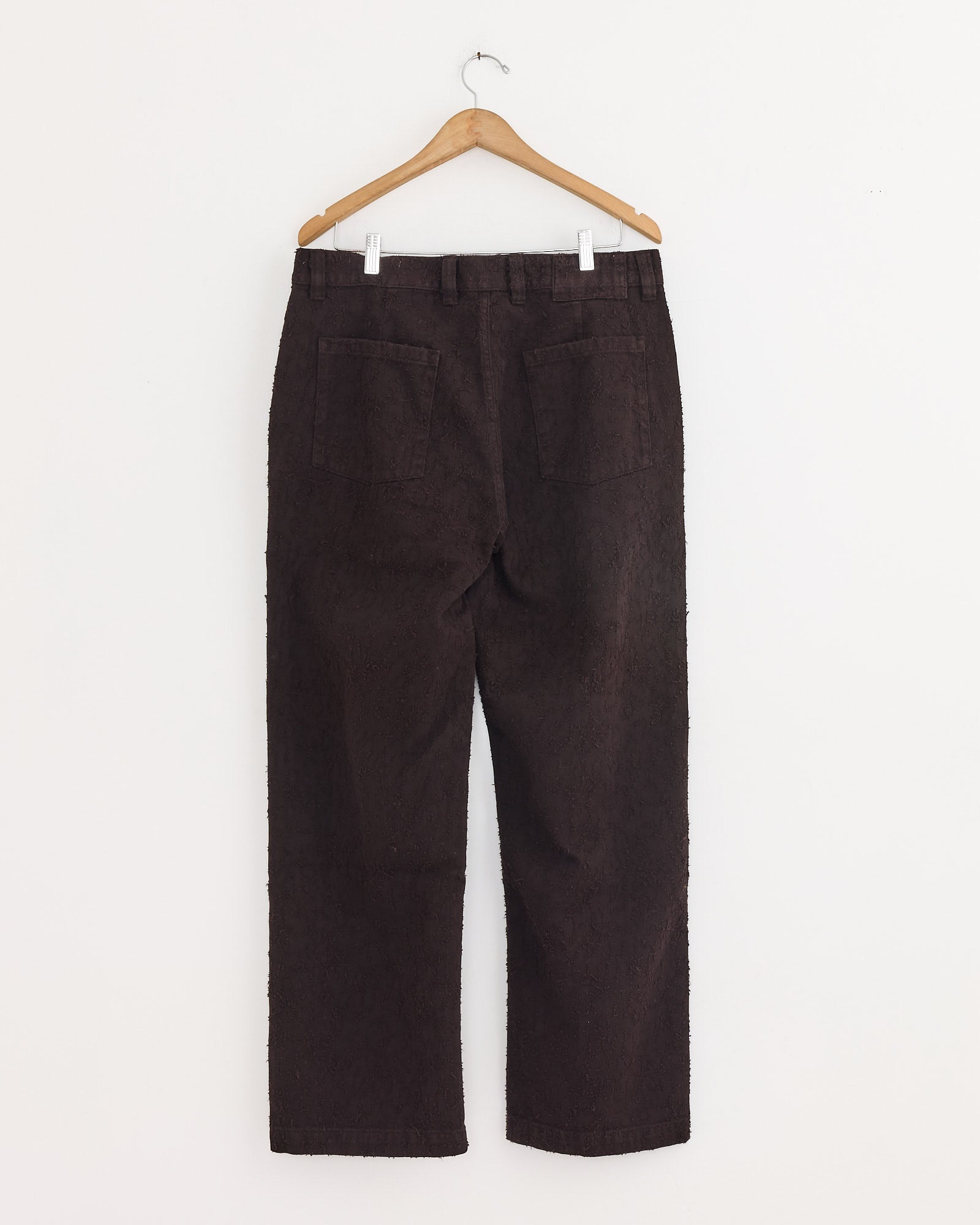 The Punch Out Jean in Dark Brown by mfpen features straight legs and two back pockets, crafted from organic cotton denim. Shown hanging on a wooden hanger against a white wall.