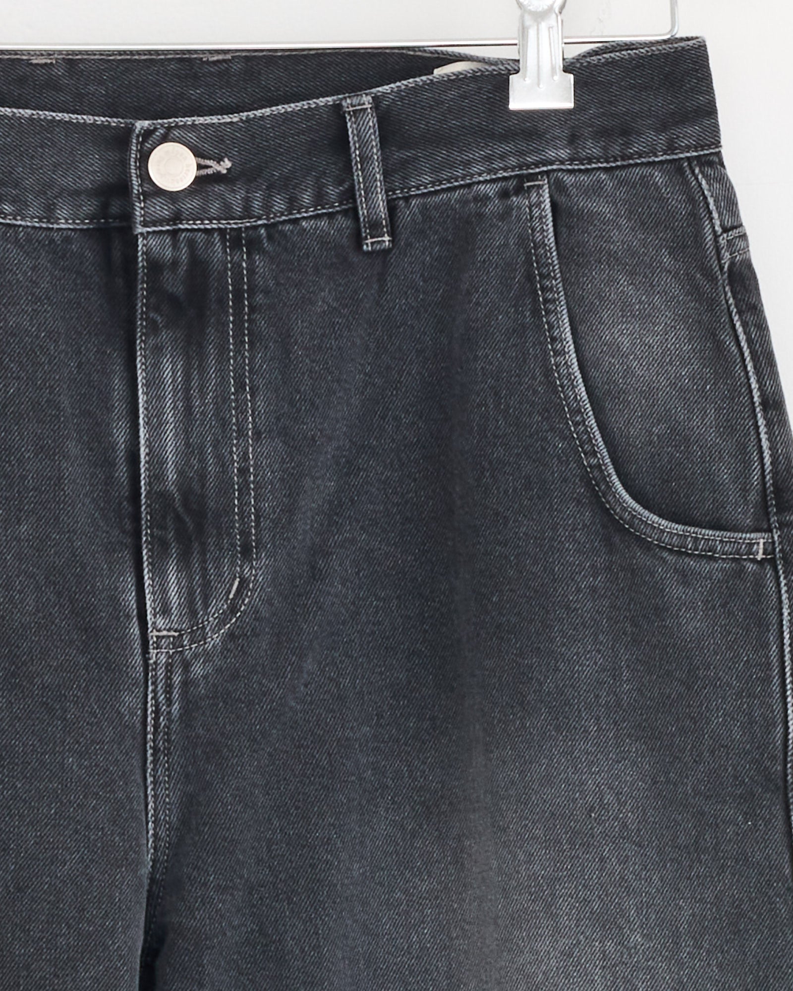 Close-up of the upper section of mfpen Regular Jean in Vintage Grey, made from organic cotton denim, hanging on a clear plastic hanger and displaying the front pocket, belt loop, and metal button closure.