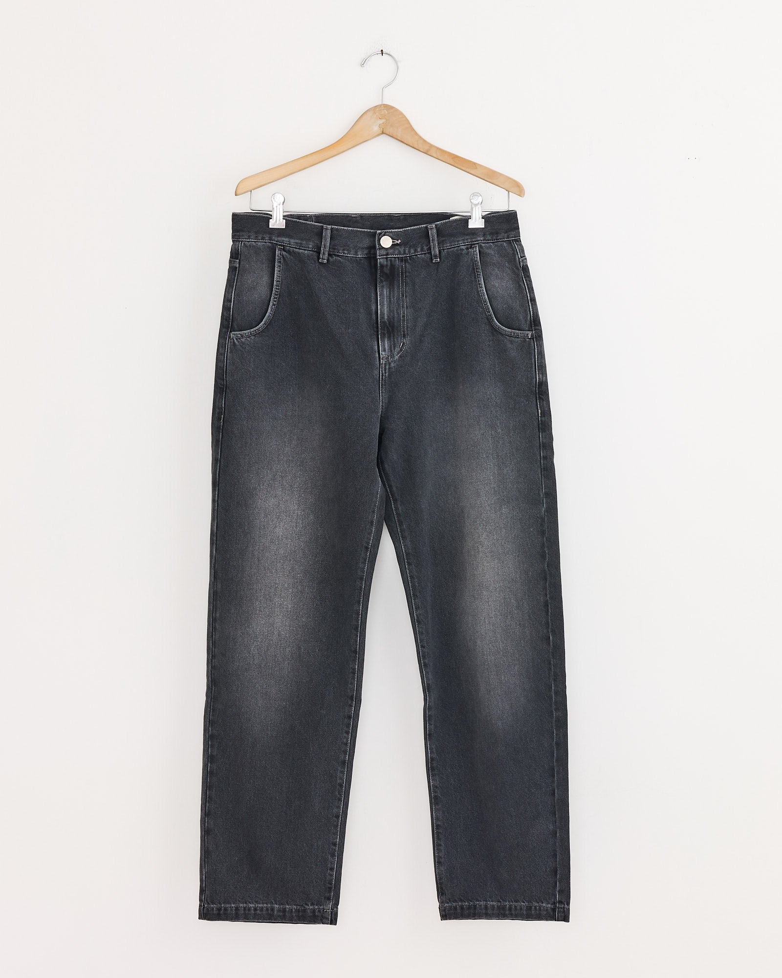 The mfpen Regular Jean in Vintage Grey, featuring a button closure, front pockets, and a straight-leg fit, hangs on a wooden hanger against a plain white wall.