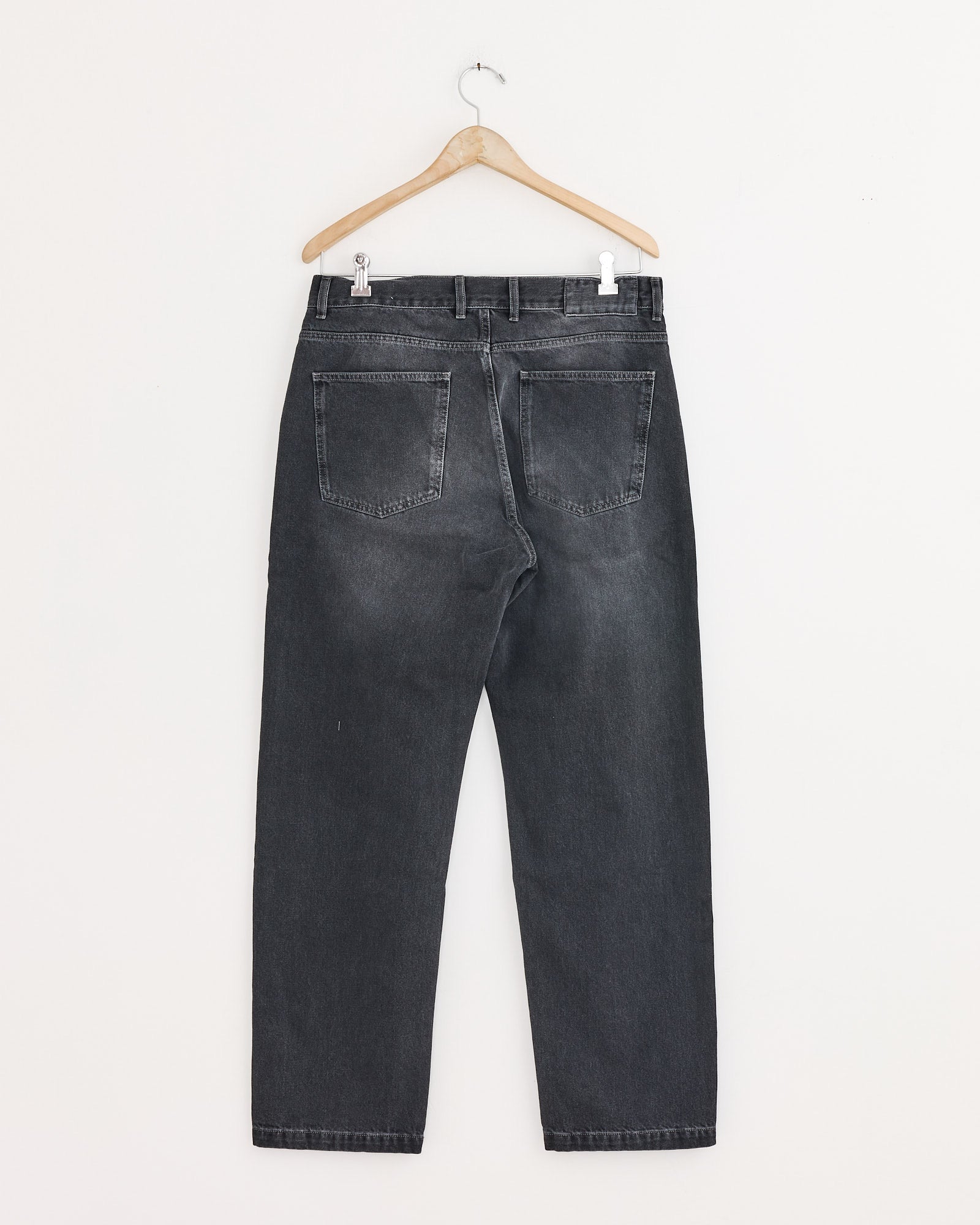 The mfpen Regular Jean in Vintage Grey, a relaxed fit style, hangs on a wooden hanger against a white wall, shown from the back with two rear pockets and belt loops visible.