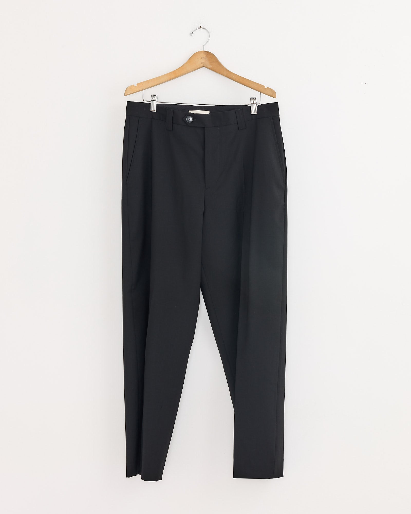 The mfpen Study Trouser in Black, a pair of wool suit pants, hangs neatly on a wooden hanger against a plain white wall.