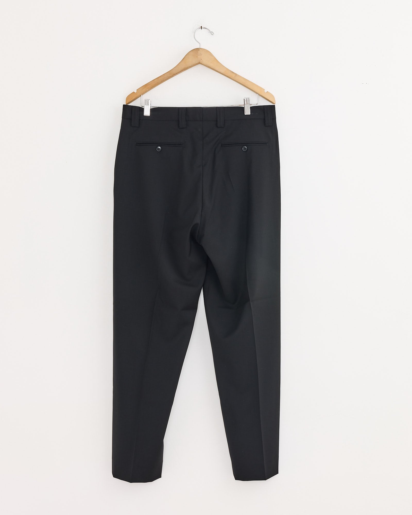The mfpen Study Trouser in Black, crafted from Italian wool, hangs on a wooden hanger against a white wall and is shown from the back with two buttoned rear pockets.