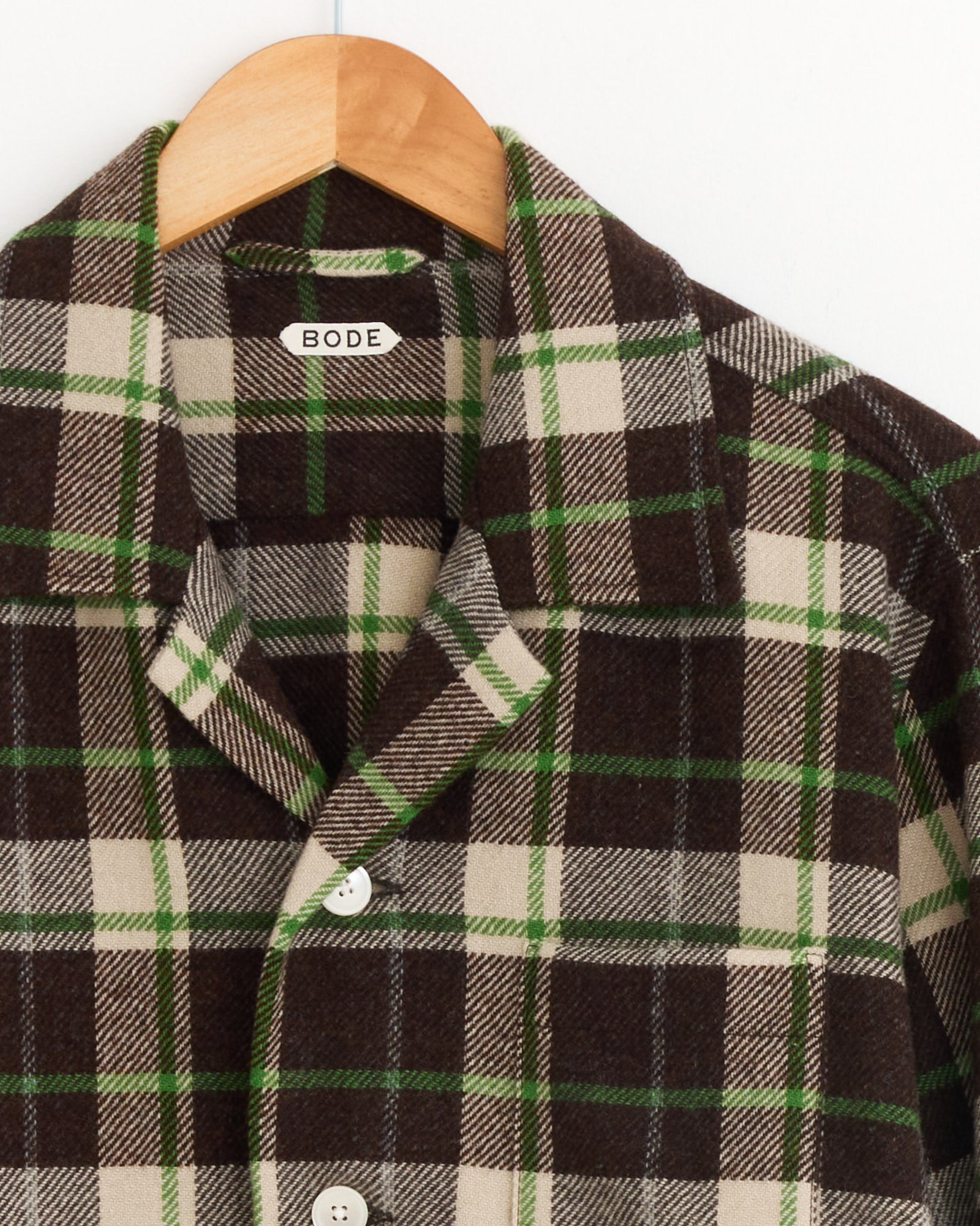 A close-up of the Bode Woodcrest Overshirt in Brown/Green, featuring a plaid flannel design with a collar, buttoned and displayed on a wooden hanger. A white label inside the collar reads "BODE.
