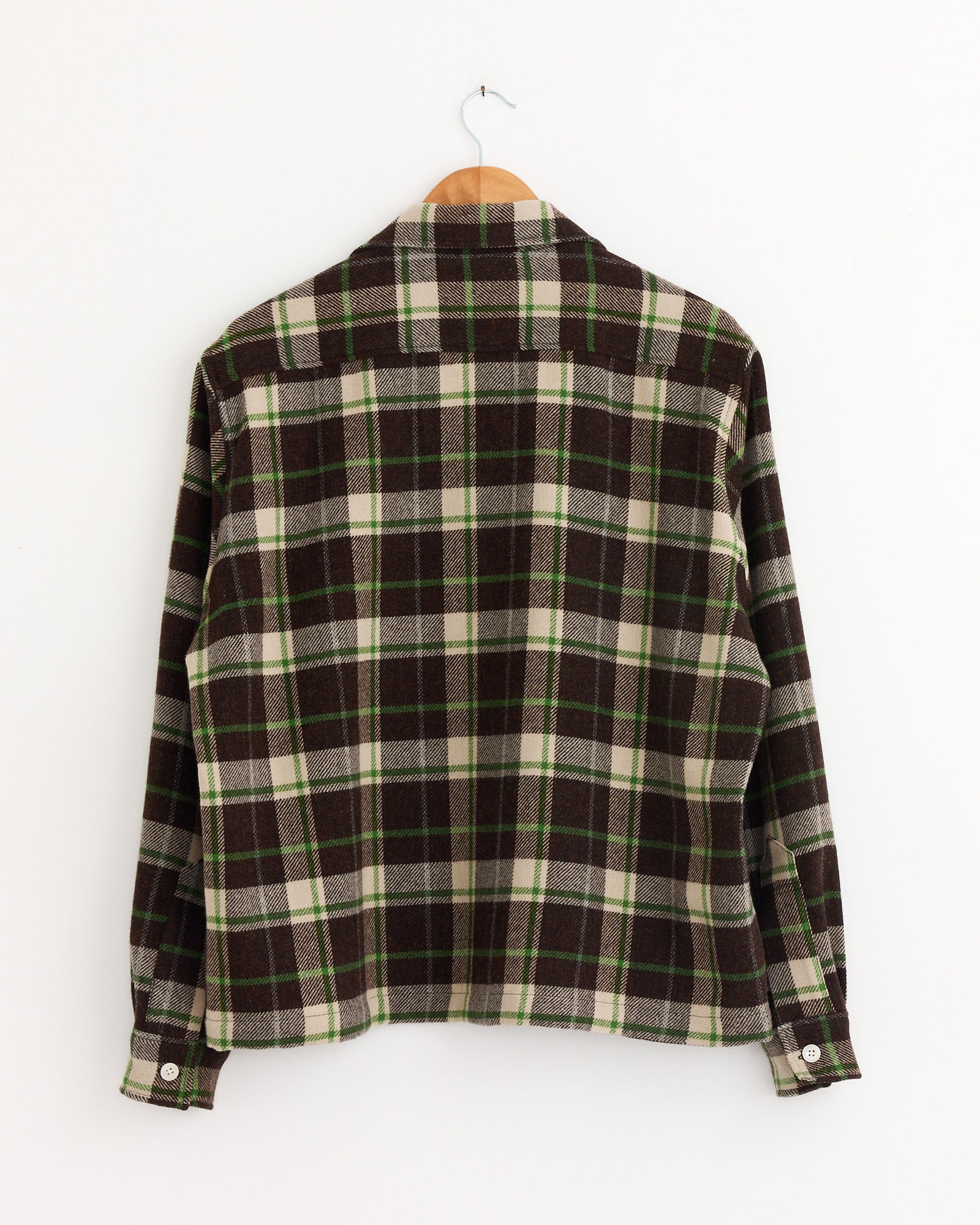 The Woodcrest Overshirt in Brown/Green by Bode features brown, green, and white plaid flannel, shown from the back on a wooden hanger against a plain white wall with sleeves slightly bent downward.