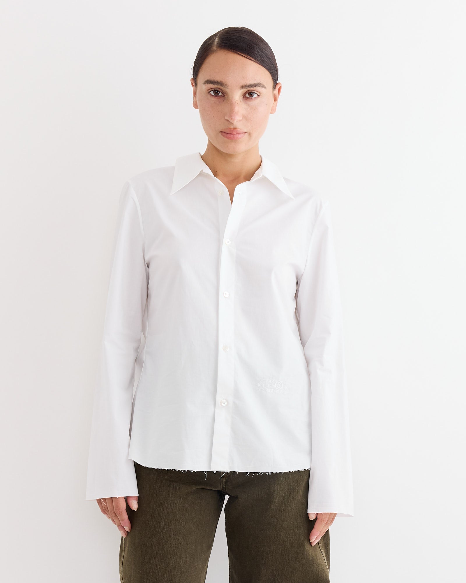 A person with dark hair parted in the middle wears an MM6 Maison Margiela Long Sleeve Shirt in White, paired with olive green pants, standing against a plain white background.
