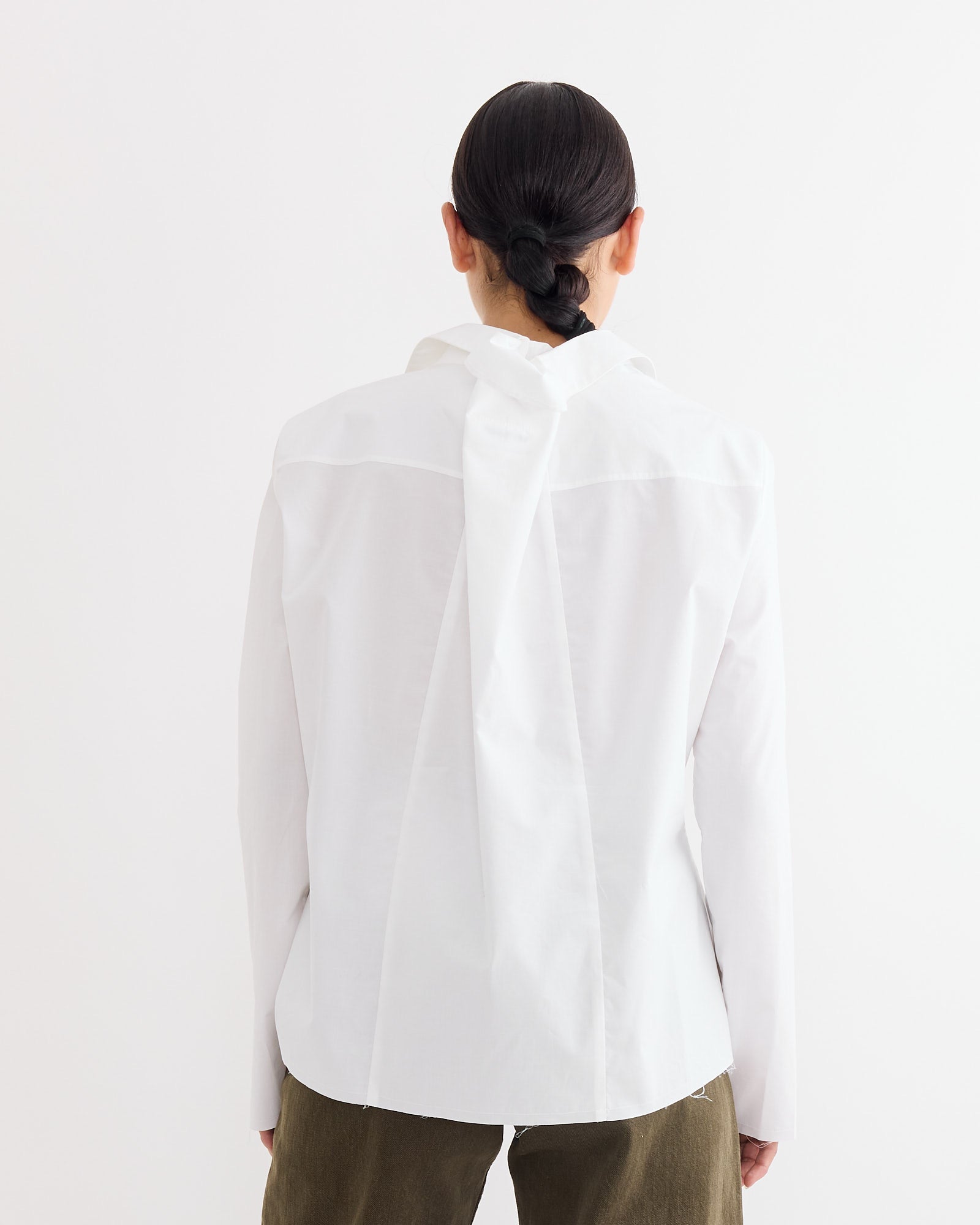 Seen from behind, a person with dark hair in a low bun wears the MM6 Maison Margiela Long Sleeve Shirt in White, featuring folded back detail and subtle Numeric Signature embroidery, paired with olive green pants against a white background.