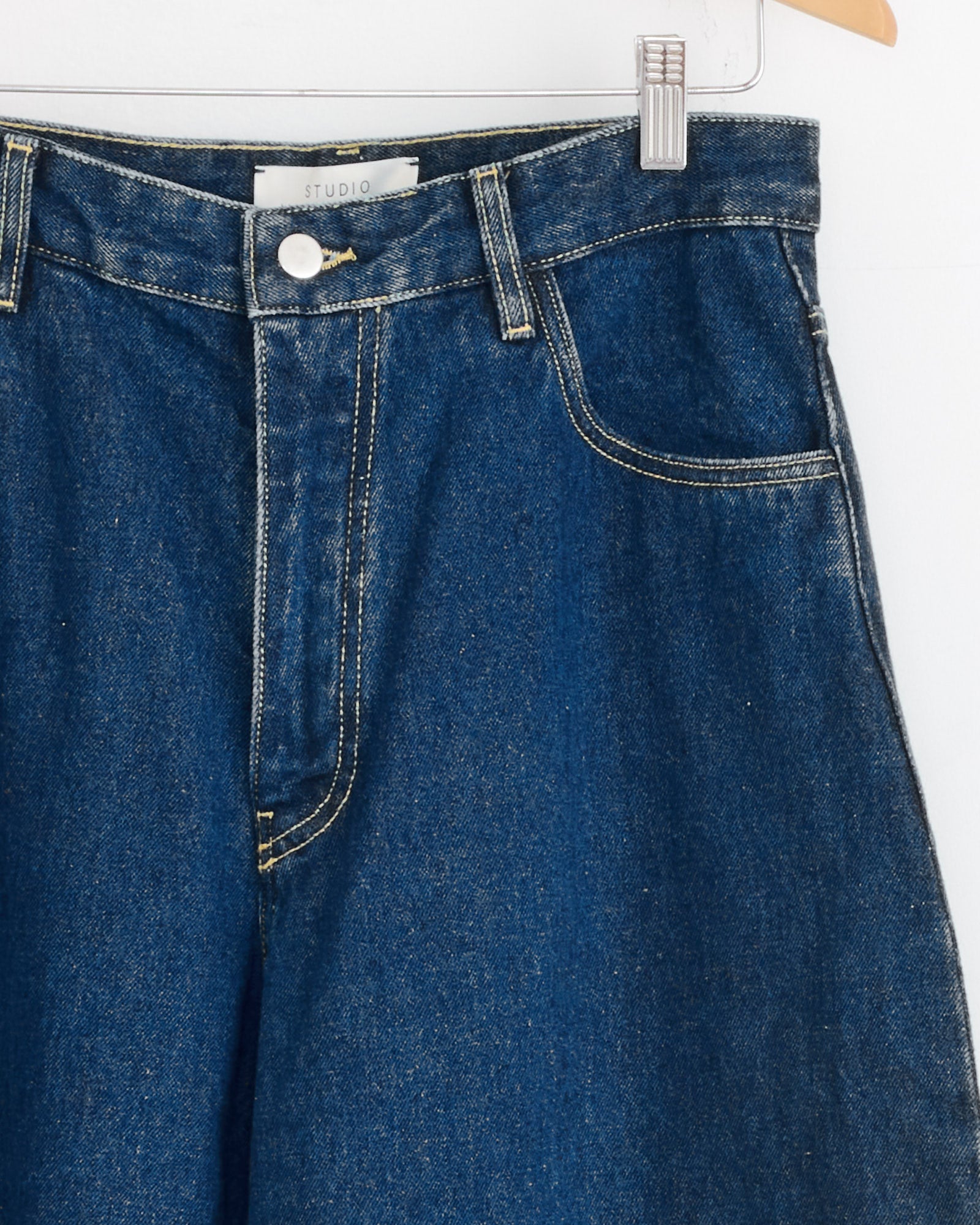 A close-up of the Paolo Pant in Indigo Wash by Studio Nicholson, hanging on a wooden hanger, highlights the waistband, button, front pockets, white Studio Nicholson label, contrast stitching, and classic design.