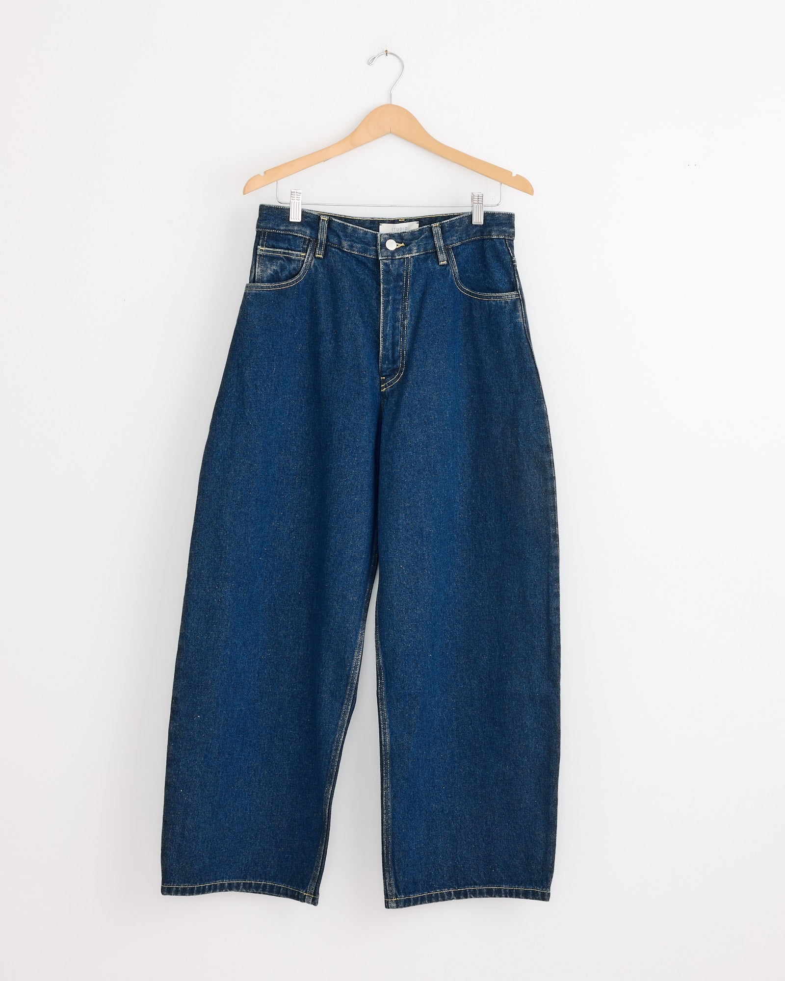 The Paolo Pant in Indigo Wash by Studio Nicholson, crafted from regenerative cotton, hangs on a wooden hanger against a plain white wall.