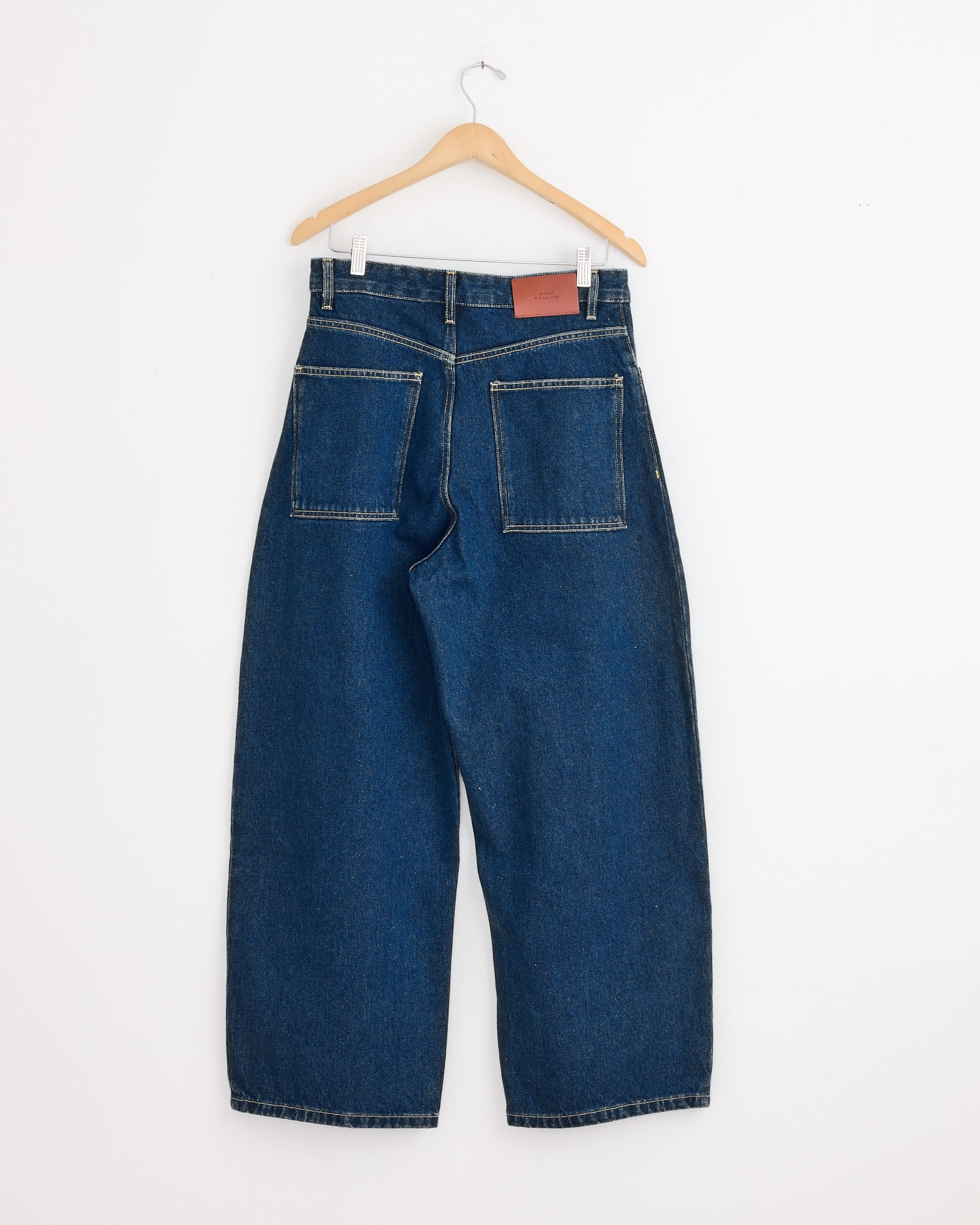 The Paolo Pant in Indigo Wash by Studio Nicholson, featuring wide-leg indigo-washed denim and large back pockets, hangs on a wooden hanger against a plain white wall.