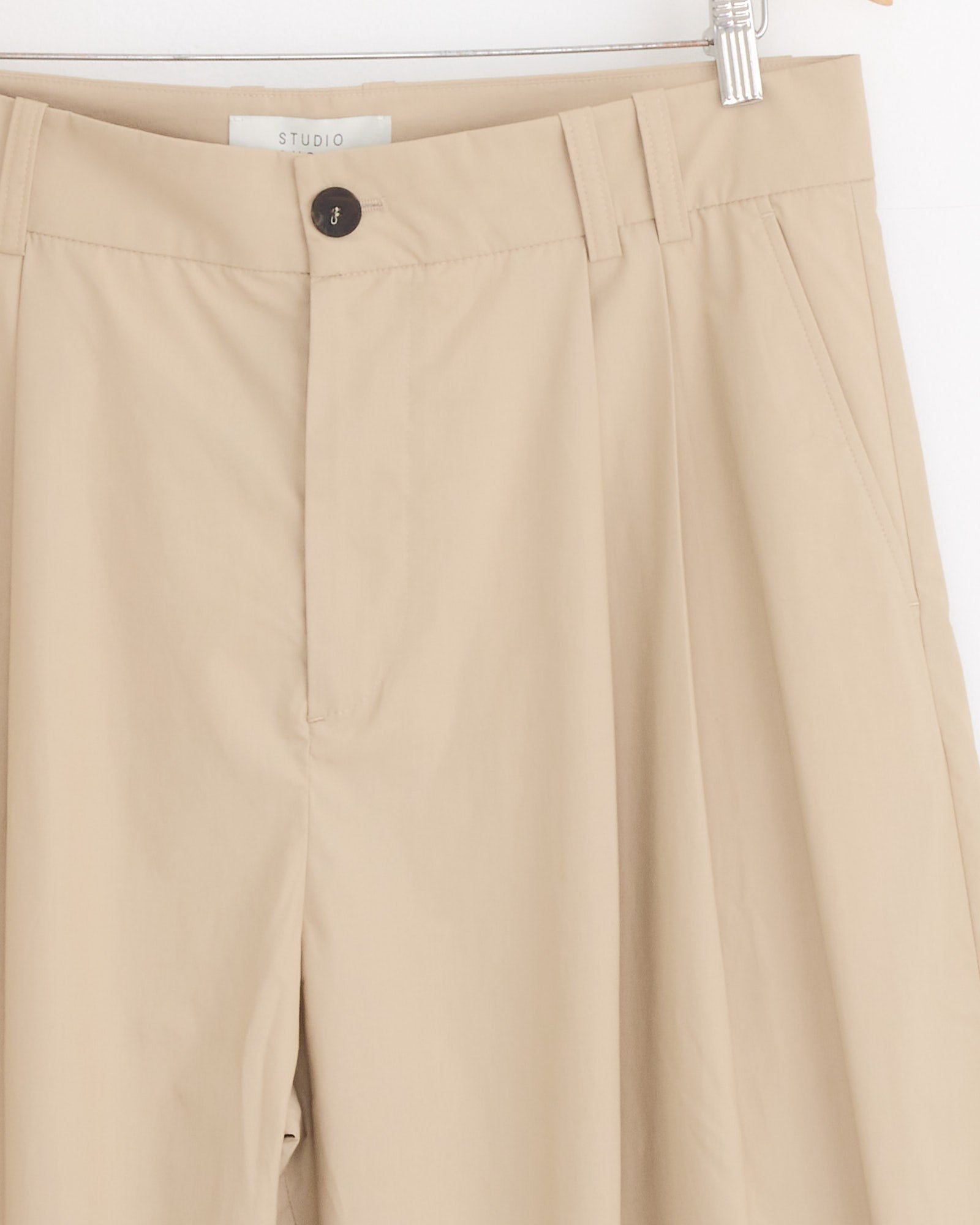 Close-up of Studio Nicholson’s Yale Pant in Reed, hanging on a hanger. Made from Italian-milled poplin, these beige trousers feature a waistband with belt loops, black button, side pockets, and a minimalist relaxed silhouette.