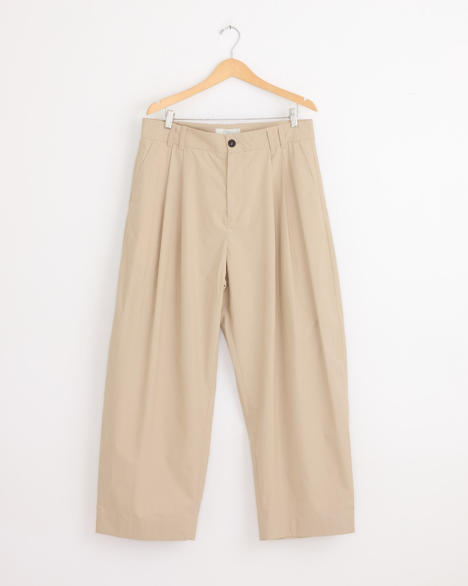 The Studio Nicholson Yale Pant in Reed, featuring a wide-leg silhouette and crafted from Italian-milled poplin with pleats and a button closure, hangs on a wooden hanger against a white backdrop.