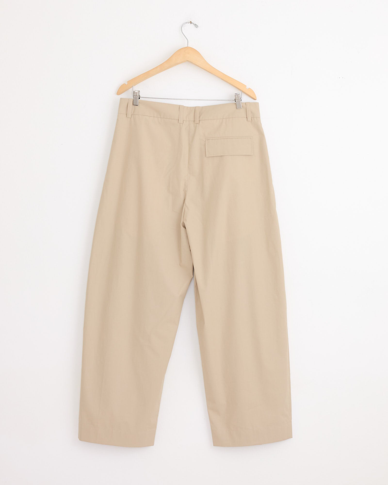 Studio Nicholson's Yale Pant in Reed, crafted from Italian-milled poplin, hangs on a wooden hanger against a white wall. The back view shows its relaxed silhouette and a single right-side back pocket.