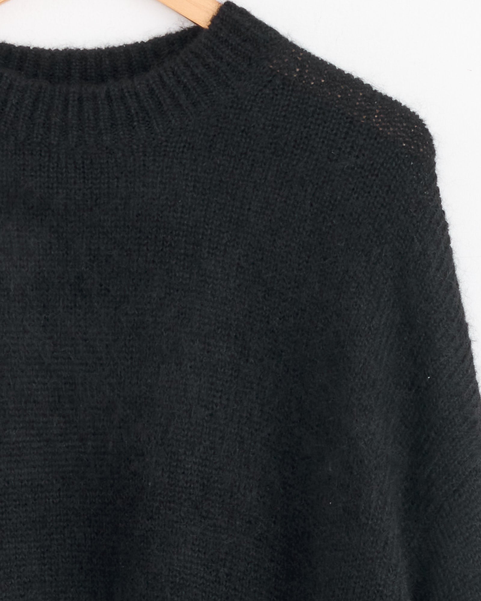 A close-up of the Studio Nicholson Wynn Knit in Black, an Italian-made superkid mohair sweater, hangs on a wooden hanger. The knit texture and ribbed collar stand out against a white background.