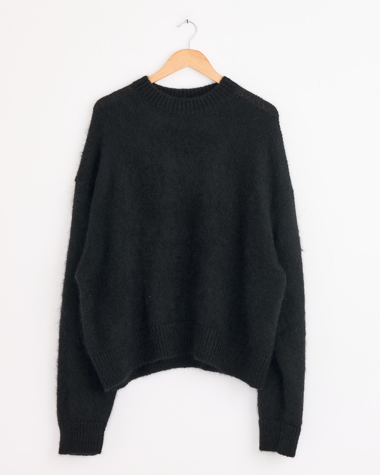 The Studio Nicholson Wynn Knit in Black hangs on a wooden hanger against a white wall. This long-sleeve sweater features a loose fit, ribbed cuffs, and a ribbed crew neckline.