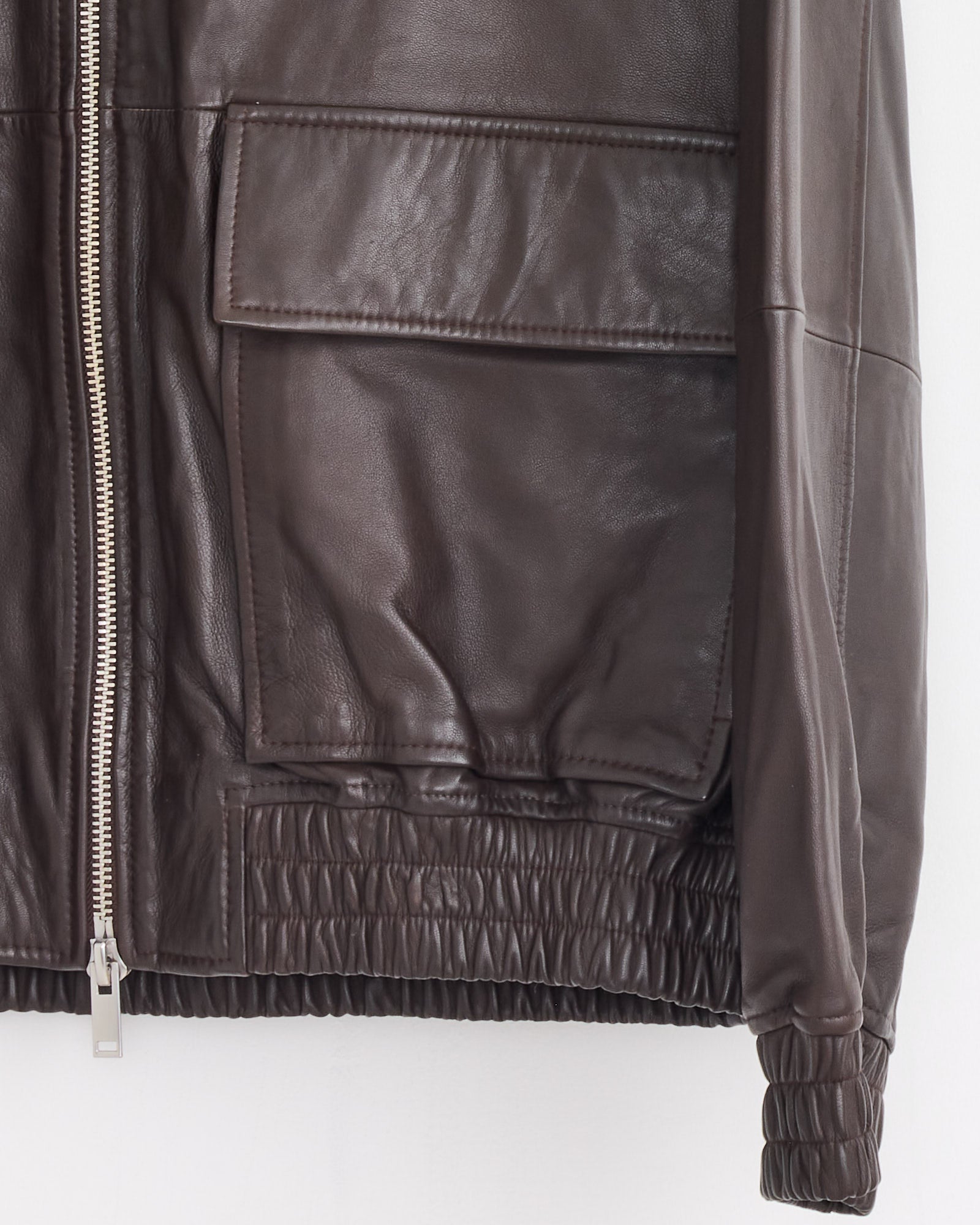 Close-up of the Studio Nicholson Piston Bomber Jacket in Brown lamb leather, featuring a large flap pocket, silver zipper, and elastic hem and cuffs, shown on a white background.