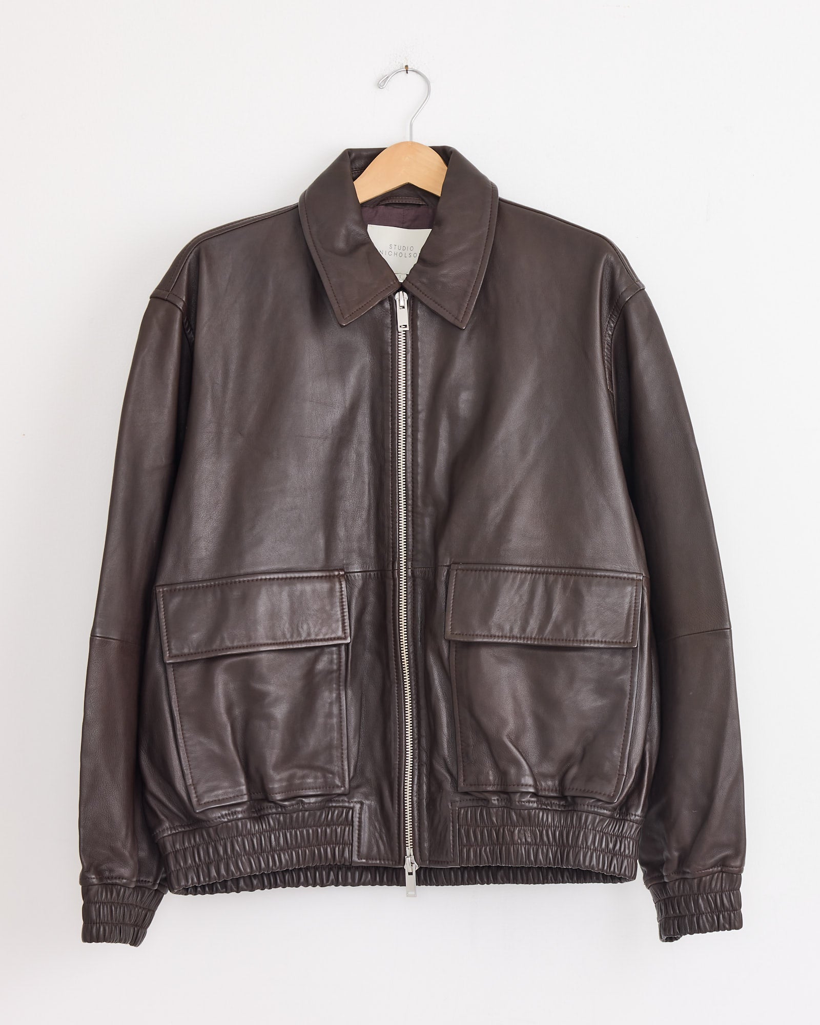 The Studio Nicholson Piston Bomber Jacket in Brown, crafted from supple lamb leather, features a zip front, two large pockets, classic collar, and elastic cuffs and hem. Shown hanging on a wooden hanger against a white wall.