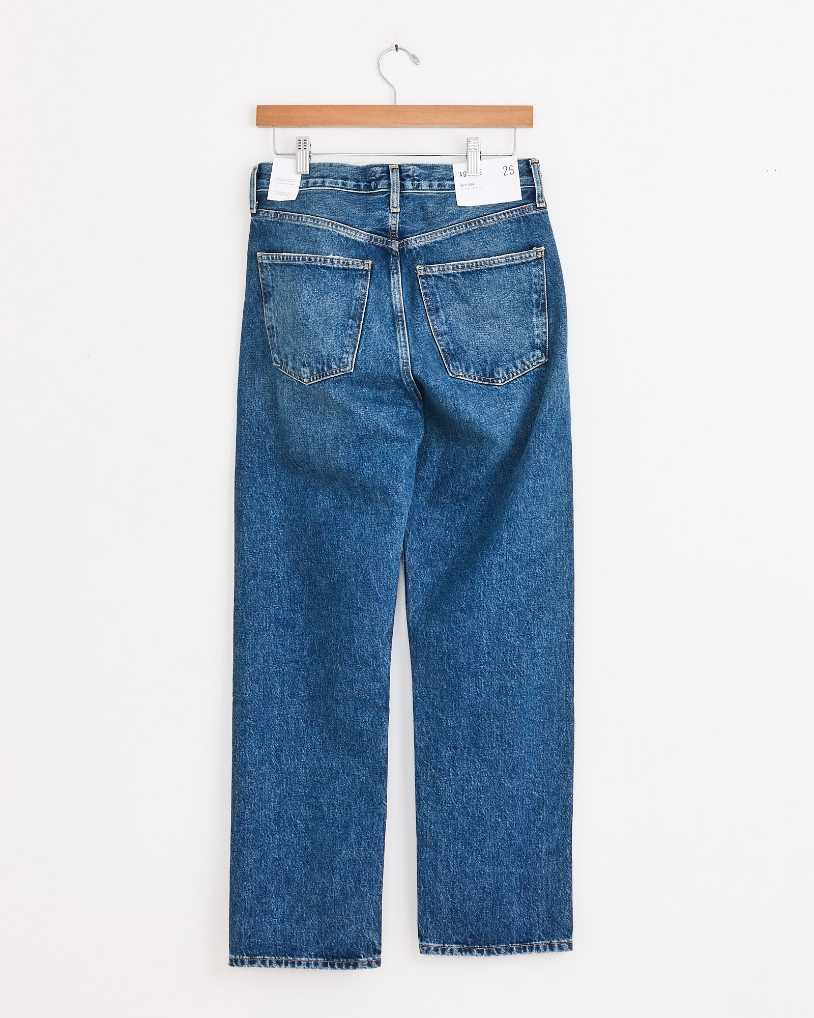 The AGOLDE 90s Jean in Bounty, made from regenerative cotton denim, hangs on a wooden hanger against a white background, displayed from the back with two pockets and straight-cut legs.