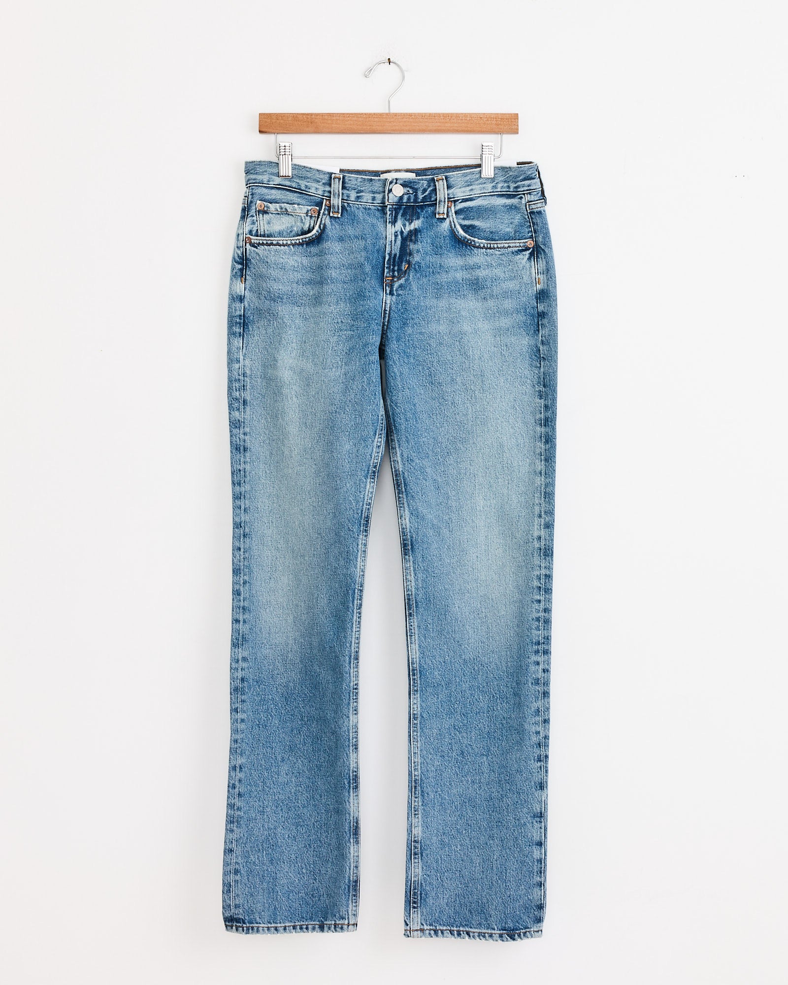 The AGOLDE Low Rise Slim in Downtown jeans feature a straight-leg fit with button and zipper closure, shown hanging on a wooden hanger against a white background. For sizing help, use our online chat.