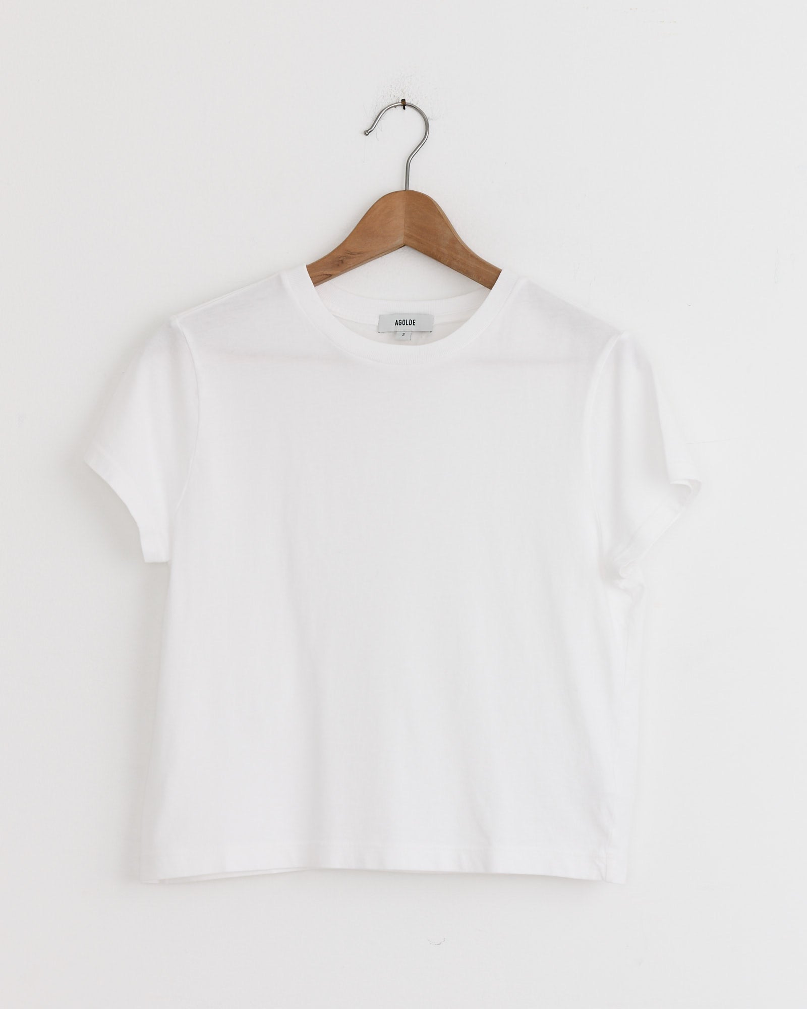 The Adine Shrunken Tee in White by AGOLDE hangs on a wooden hanger against a white wall—a versatile pick for any fit. Chat with us online for sizing advice.