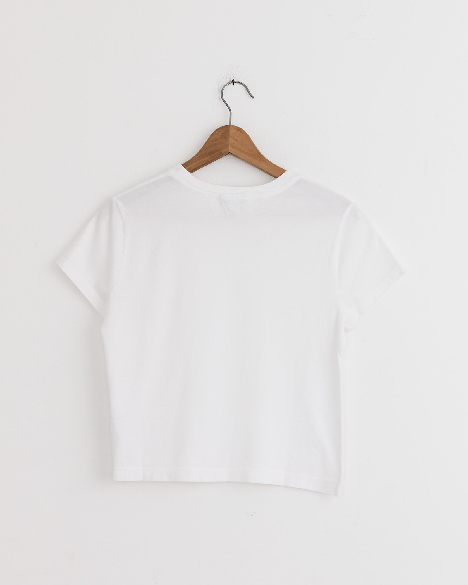 The AGOLDE Adine Shrunken Tee in White is displayed from the back on a wooden hanger against a white wall. For fit or sizing questions, our online chat support can help you find your perfect tee.