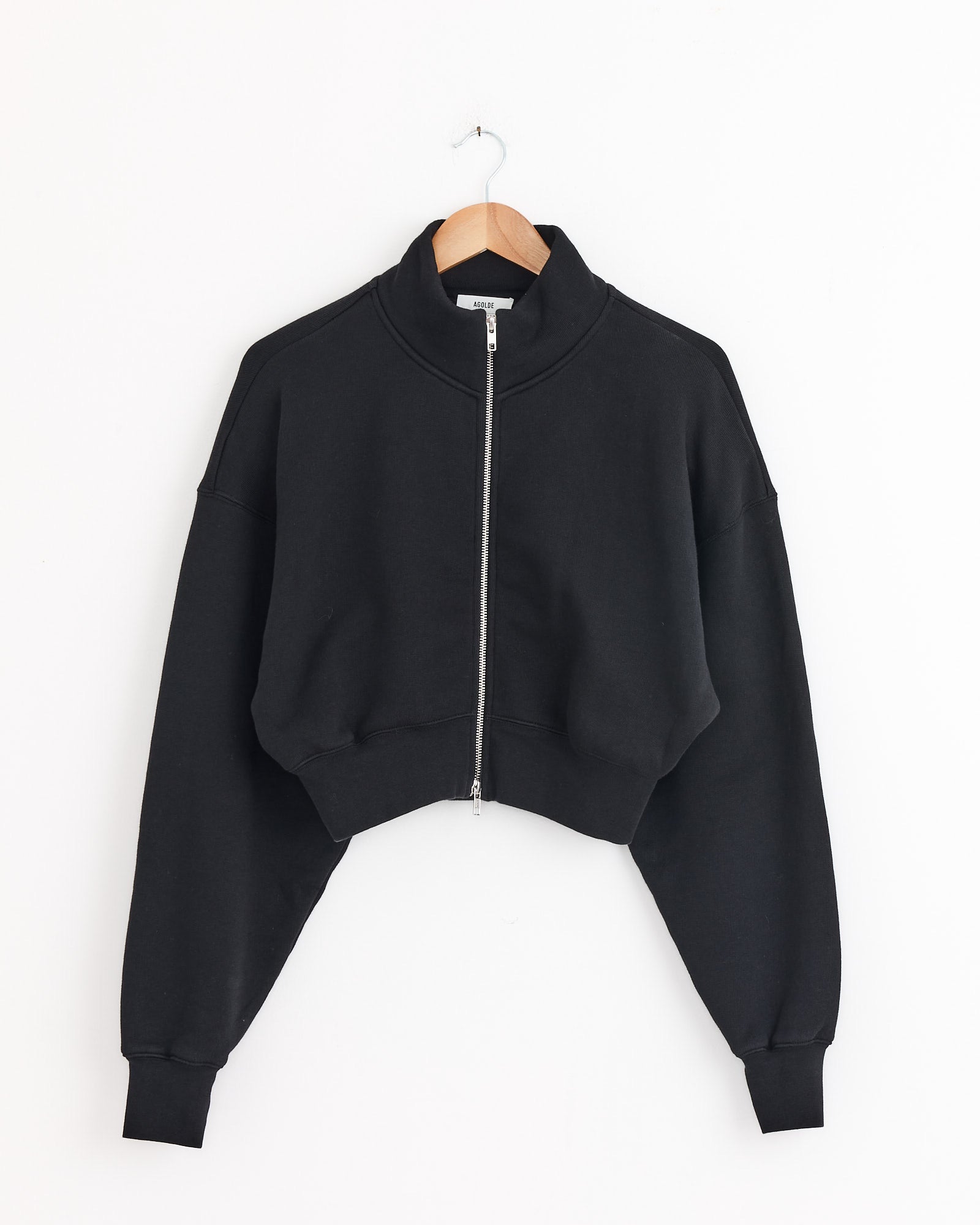 The Asta Zip Sweatshirt in Washed Black by AGOLDE, featuring long sleeves, a high collar, and cropped zip-up design, hangs on a wooden hanger against a white background. For fit or sizing questions, contact our online chat.
