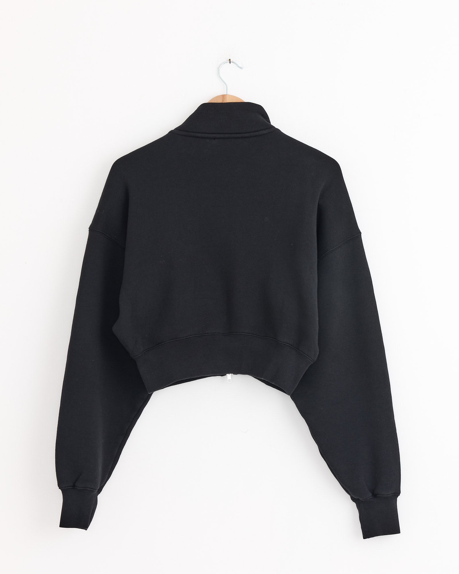 The AGOLDE Asta Zip Sweatshirt in Washed Black, a long-sleeve cropped style, is shown back-facing on a wooden hanger against a white wall. For fit or sizing questions, contact us via our online chat.