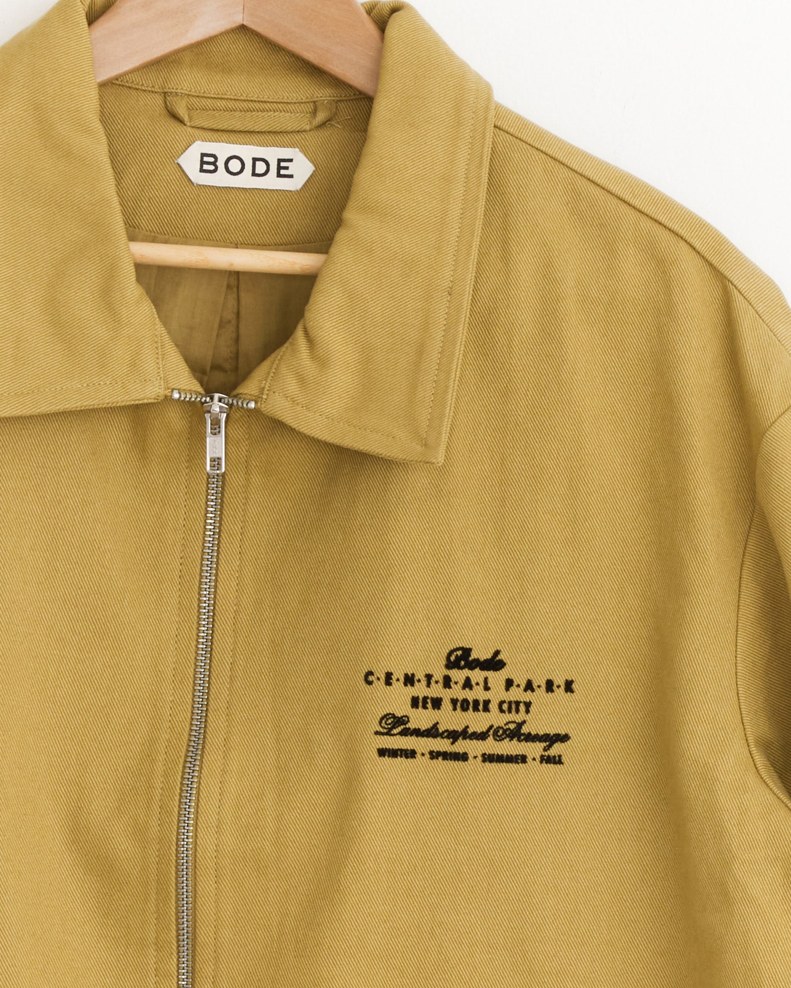 An ocre Bode Central Park Jacket hangs on a wooden hanger, featuring a front zipper and bold black embroidered "C.E.N.T.R.A.L. P.A.R.K NEW YORK CITY" text, with additional smaller words below.