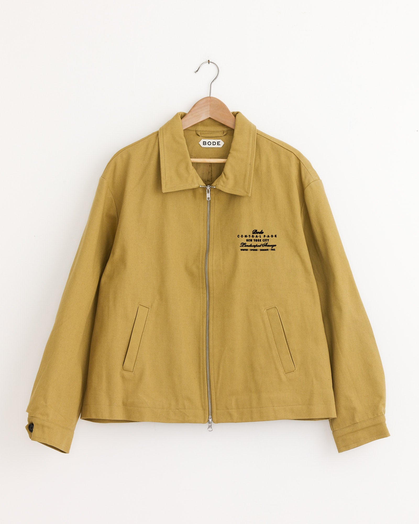 A Bode Central Park Jacket in Ocre hangs on a wooden hanger, featuring a zip front, two slanted pockets, shirt-style collar, and black embroidered text on the left chest. The Bode label is visible inside the collar.