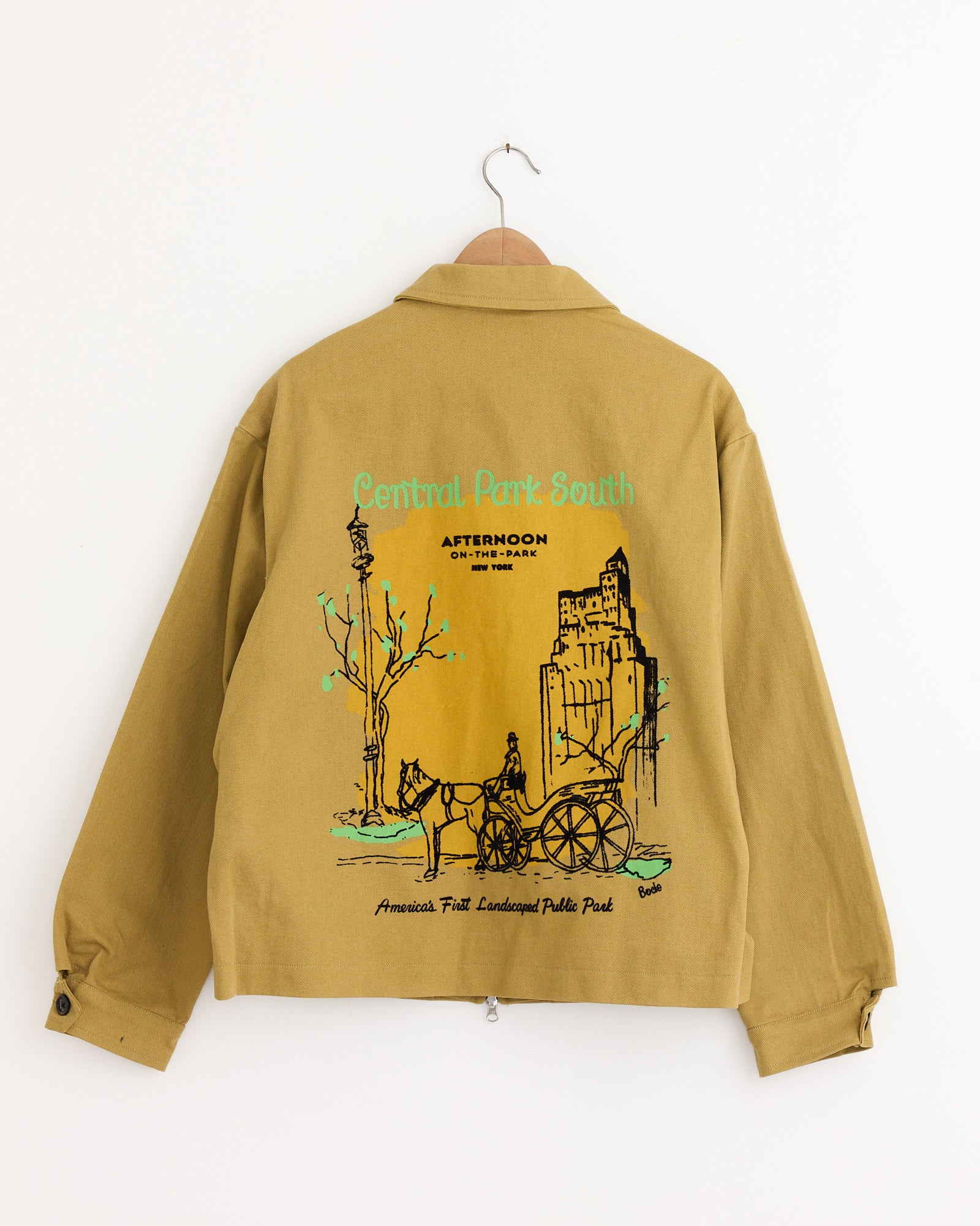 A Central Park Jacket in Ocre by Bode hangs on a wooden hanger, featuring a colorful illustration on the back of Central Park South with a horse-drawn carriage, buildings, trees, and text about Central Park.