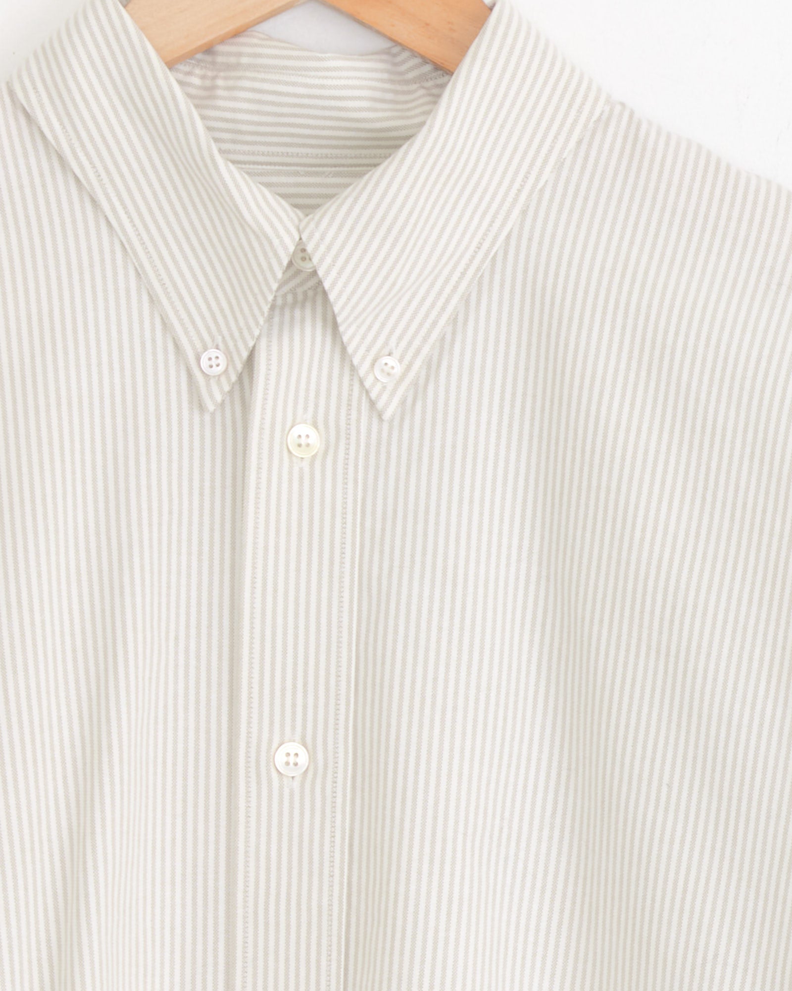 Close-up of the Studio Nicholson Ruskin Shirt in Khaki Stripe, a light beige Oxford button-down with thin vertical khaki stripes, white buttons, and a button-down collar, displayed on a wooden hanger.