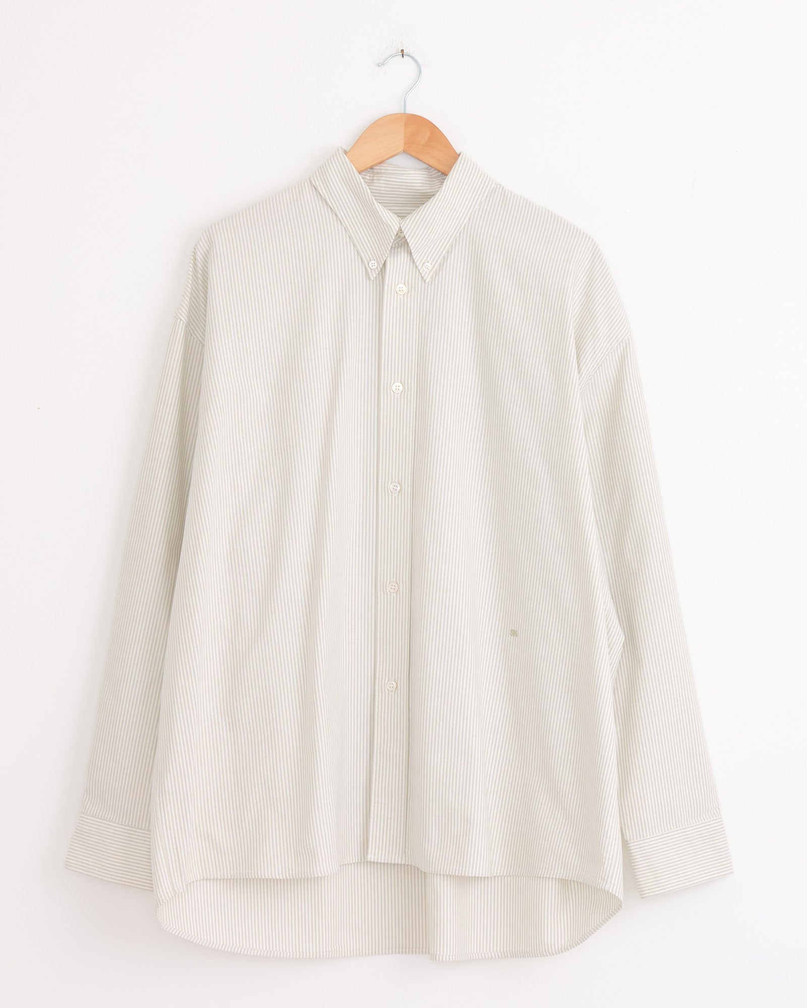 The Studio Nicholson Ruskin Shirt in Khaki Stripe, a long-sleeve light beige shirt with subtle vertical stripes, hangs on a wooden hanger against a white wall.