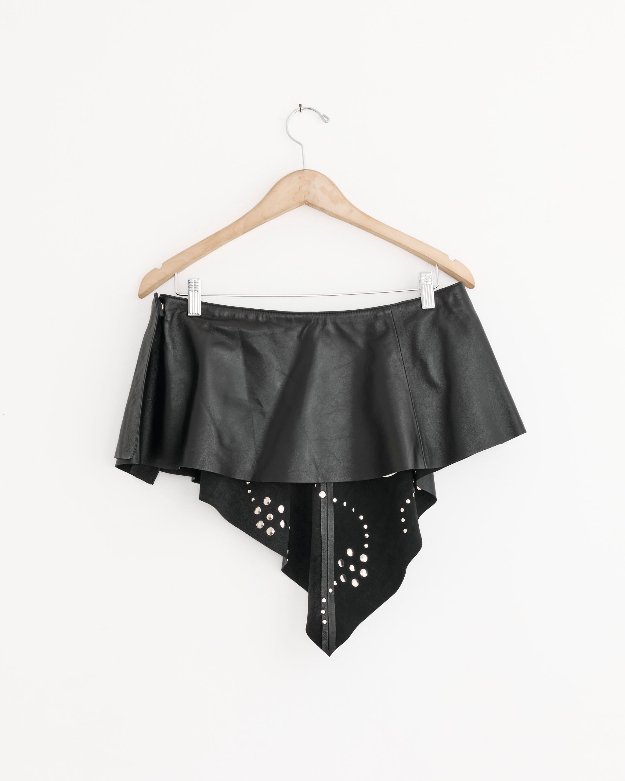 The Gimaguas Bandana Mini Skirt, a black asymmetrical leather skirt with silver studs and layered panels, is displayed on a wooden hanger against a plain white background. Available in various sizes.