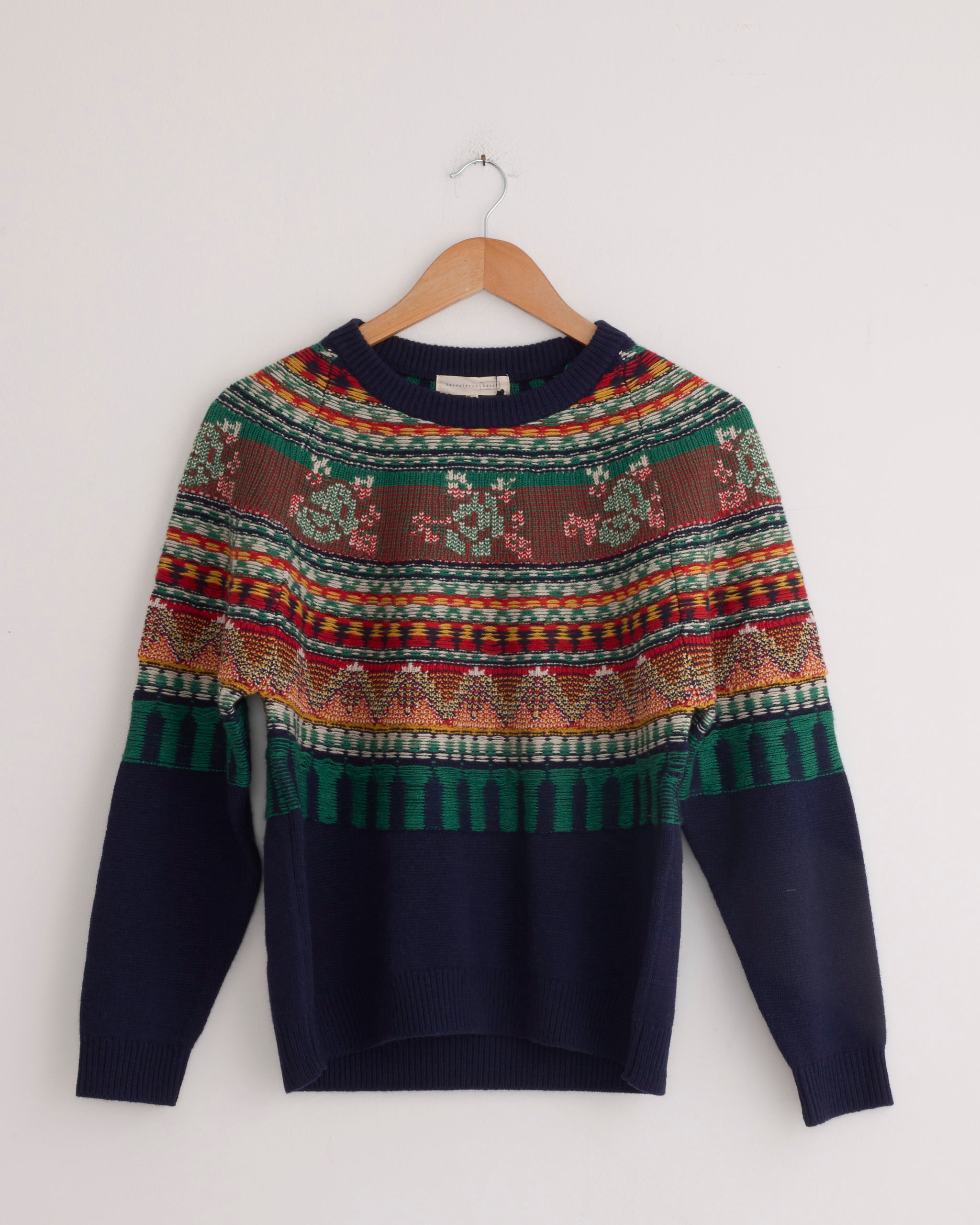 The Hansel From Basel Mabel Fairisle Pullover in Navy features vibrant knit patterns and hangs on a wooden hanger against a white wall. Use our online chat for sizing and fit assistance.