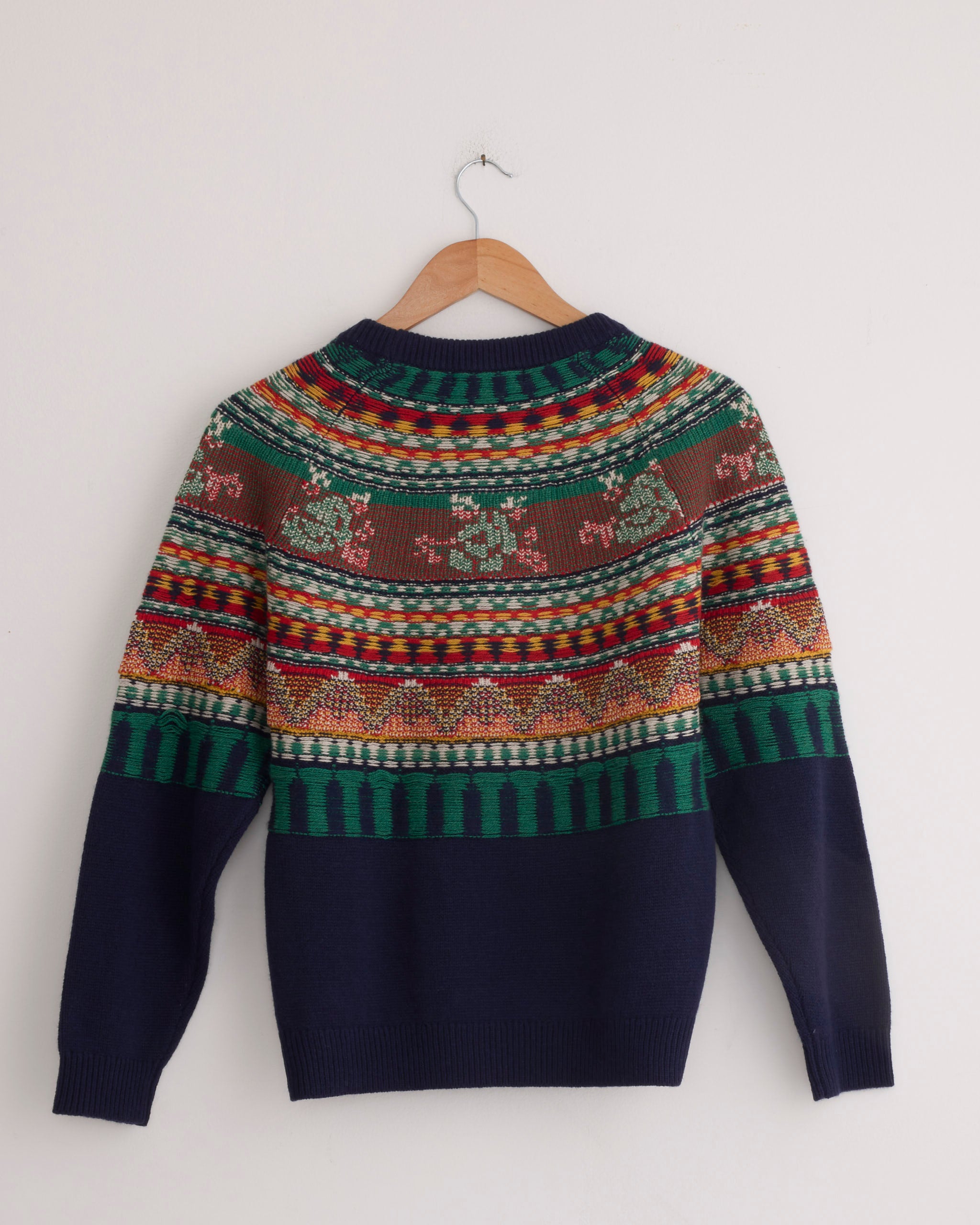 The Mabel Fairisle Pullover in Navy by Hansel From Basel features colorful geometric and floral knit patterns. Displayed on a wooden hanger against a white wall. For sizing or fit questions, our online chat can assist you.