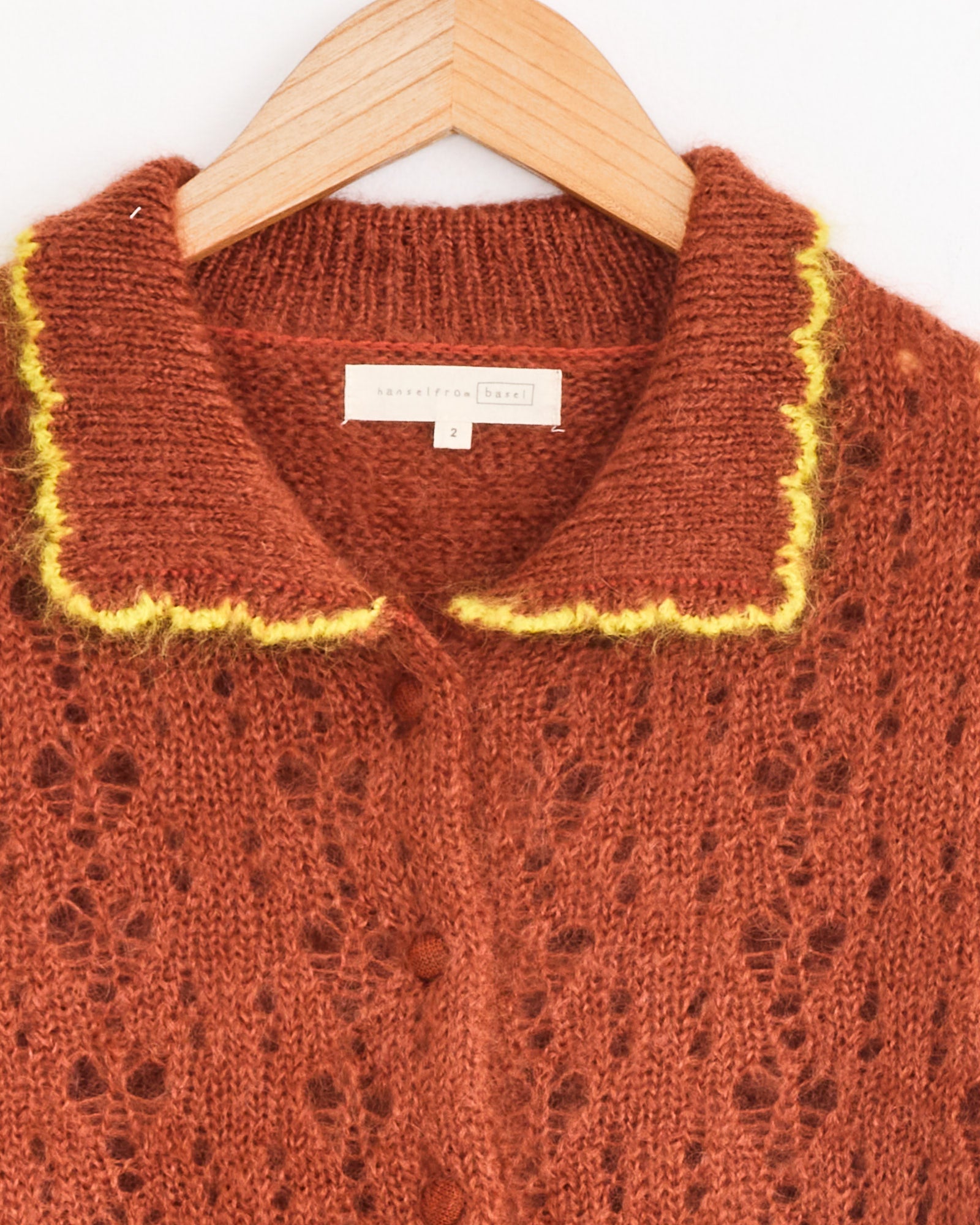 A close-up of the Hansel From Basel Ahmi Cardigan in Terracotta shows its pointelle knit, yellow-trimmed crochet collar, button front on a wooden hanger, and a white label with text and the number 2 sewn inside the neckline.