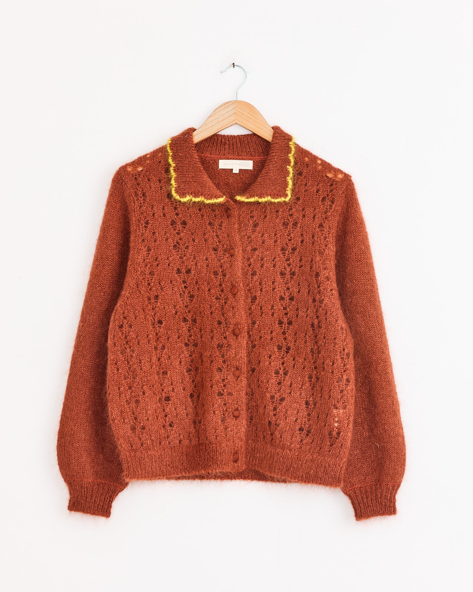 The Hansel From Basel Ahmi Cardigan in Terracotta features long sleeves, pointelle knit, ribbed cuffs and hem, yellow collar trim, and is shown on a wooden hanger against a white background.