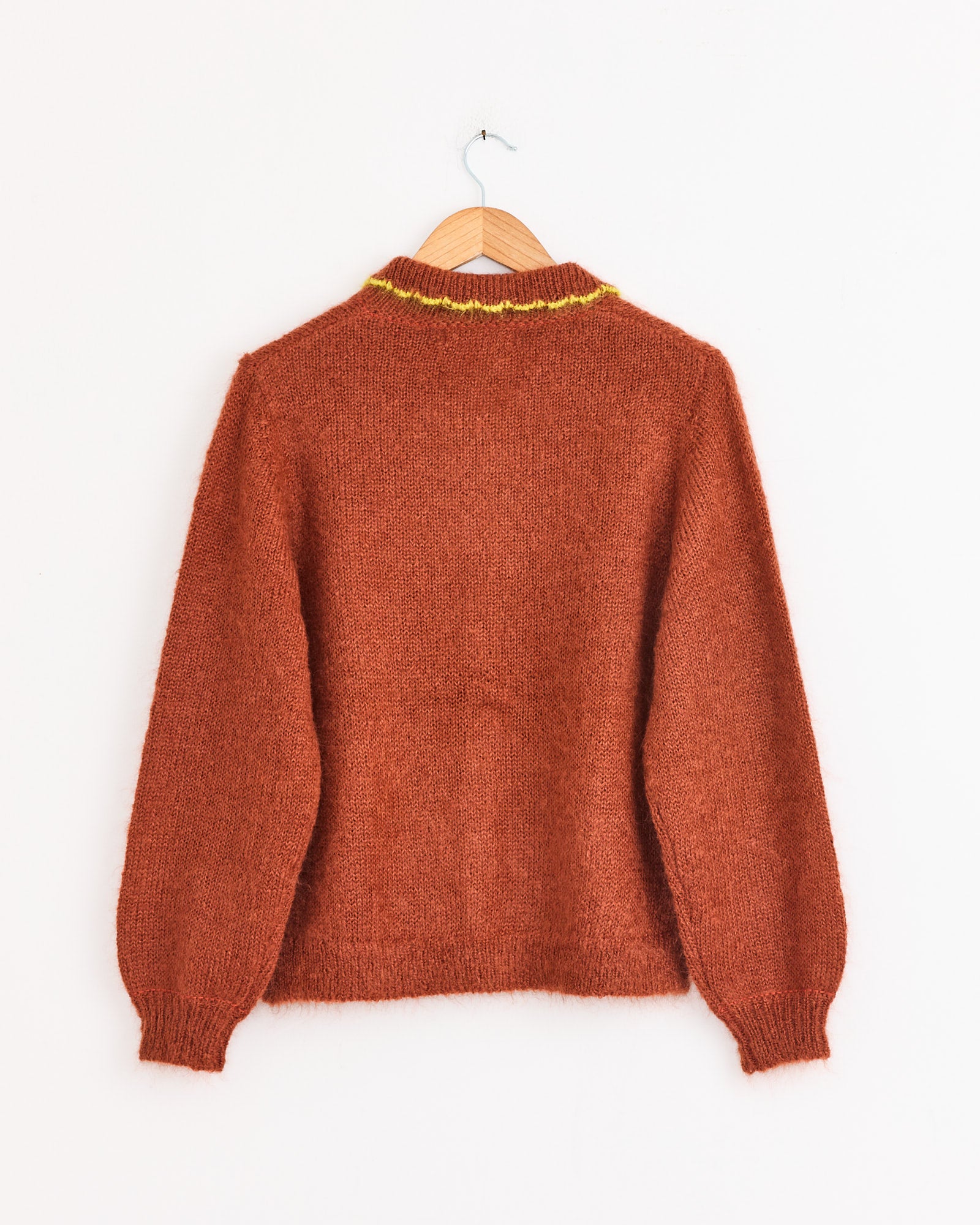 The Hansel From Basel Ahmi Cardigan in Terracotta features long sleeves, ribbed cuffs and hem, a crochet collar with a yellow accent, and textured pointelle knit. Shown on a wooden hanger against a plain white wall.
