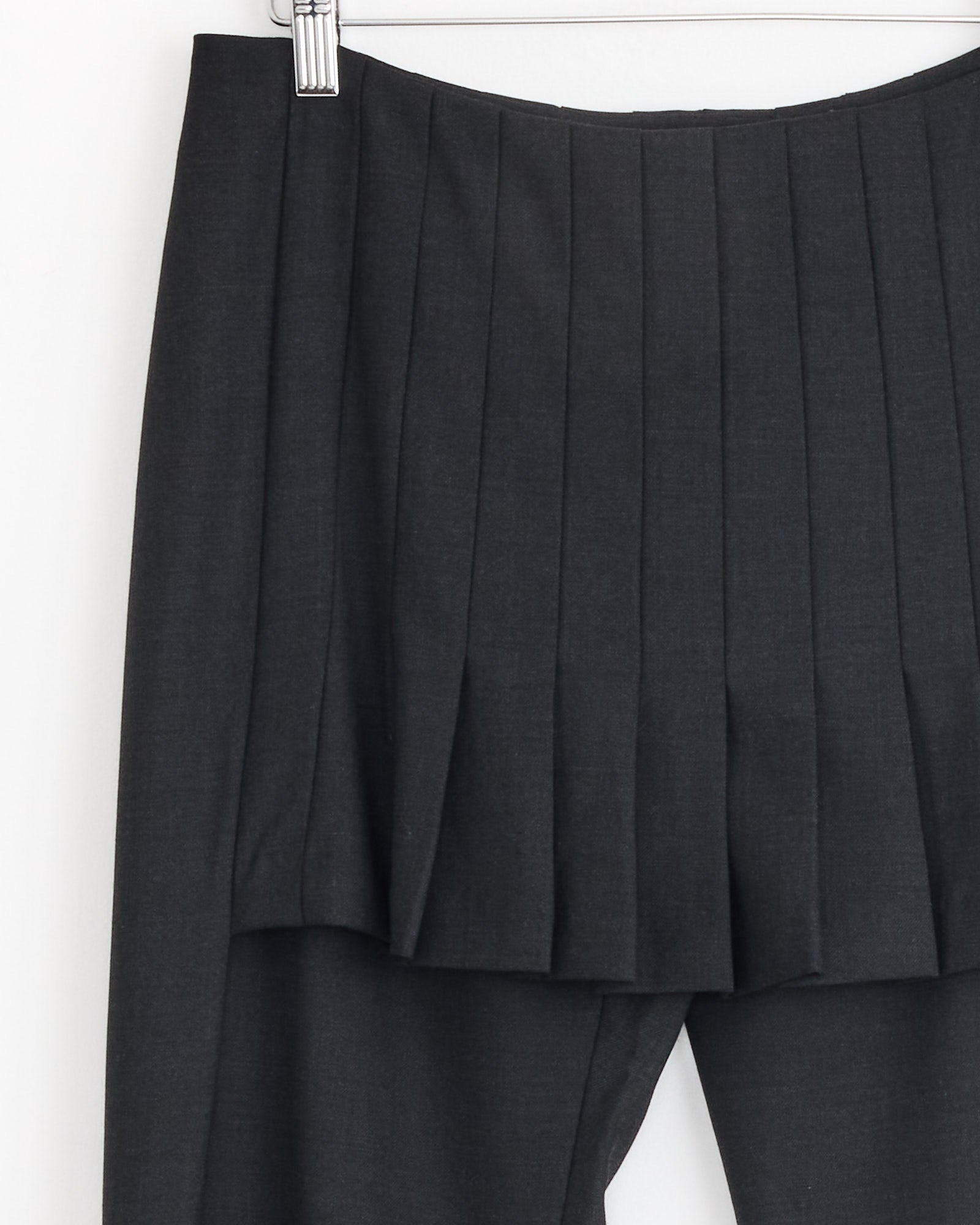 A close-up of the Paloma Wool Andrale Pant in Grey hanging on a hanger against a white background highlights its neatly pressed pleats and straight-leg, tailored silhouette.