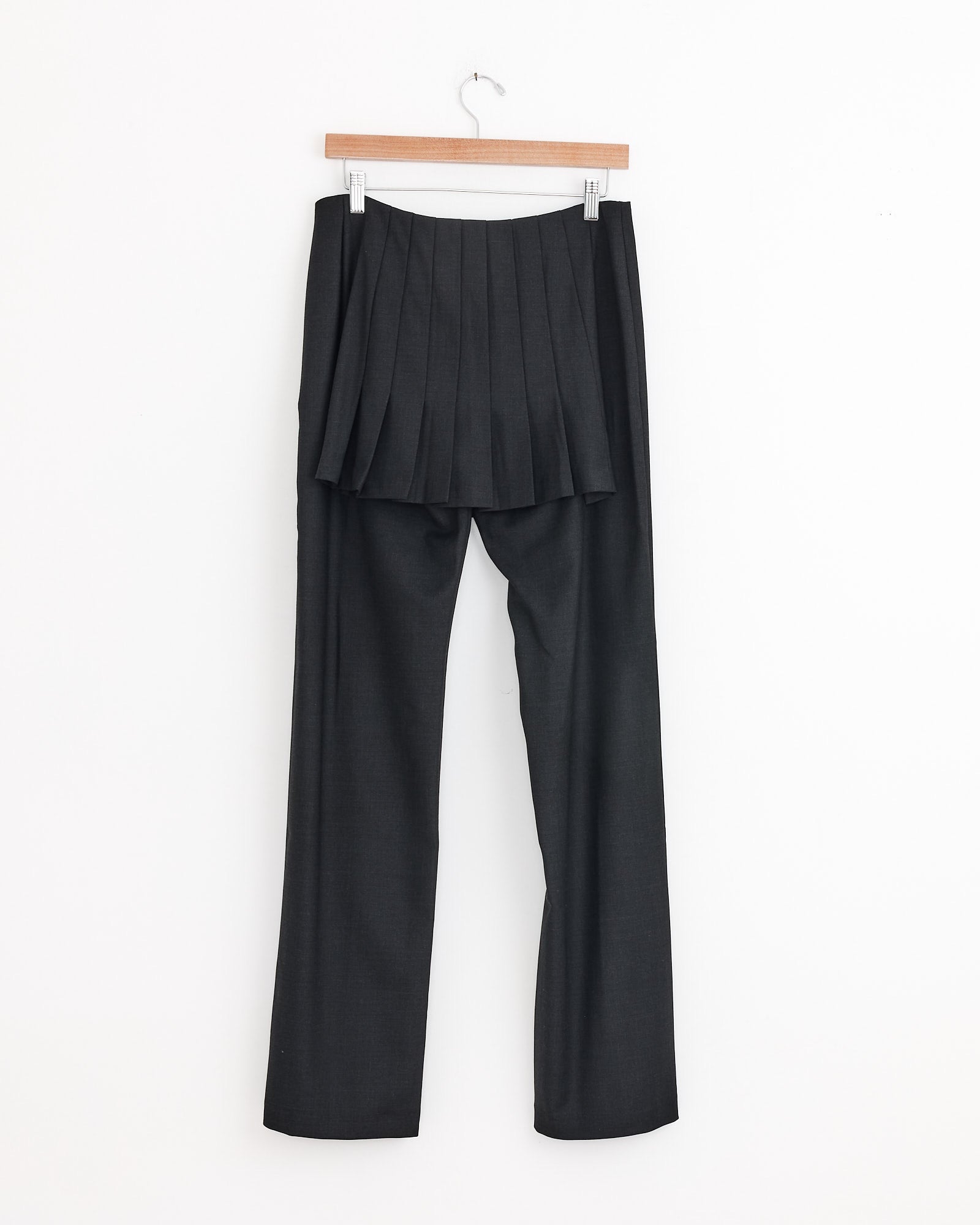A pair of Paloma Wool Andrale Pant in Grey, featuring a pleated skirt panel at the waist and a straight-leg silhouette, hangs on a wooden hanger against a white background.