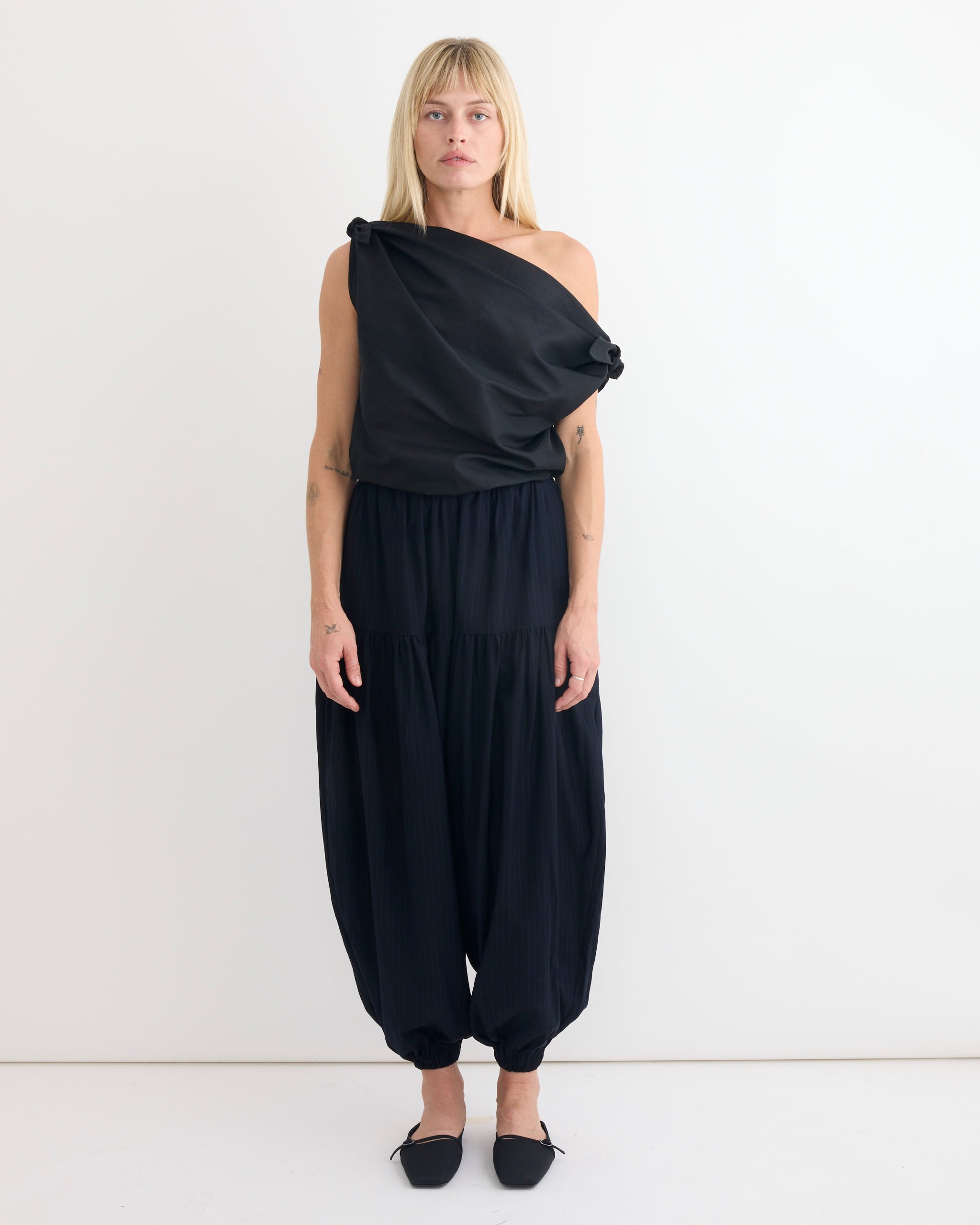 A person with straight blonde hair and a neutral expression stands against a plain white wall, wearing the Chelsea Mak Swan Top in Black paired with loose black harem pants and black flat shoes.
