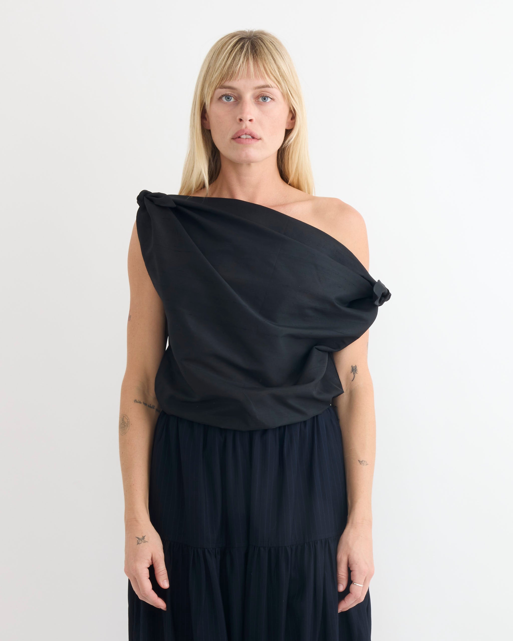 A woman with straight blonde hair wears the Chelsea Mak Swan Top in Black and matching skirt, standing against a plain white background and looking directly at the camera with a neutral expression.