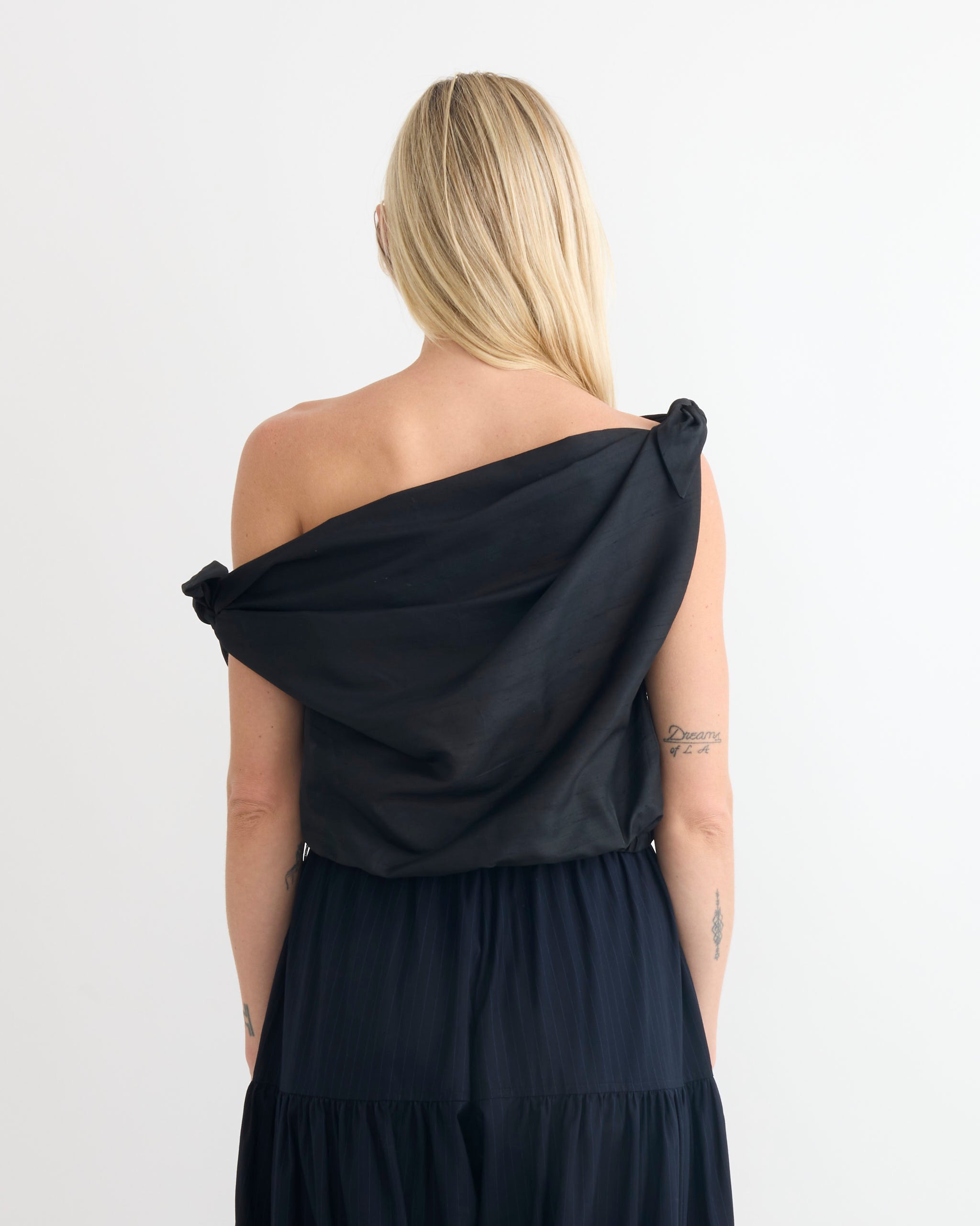 A woman with long blonde hair faces away, wearing the Chelsea Mak Swan Top in Black and a matching skirt. Small upper arm tattoos are visible against a plain white background.