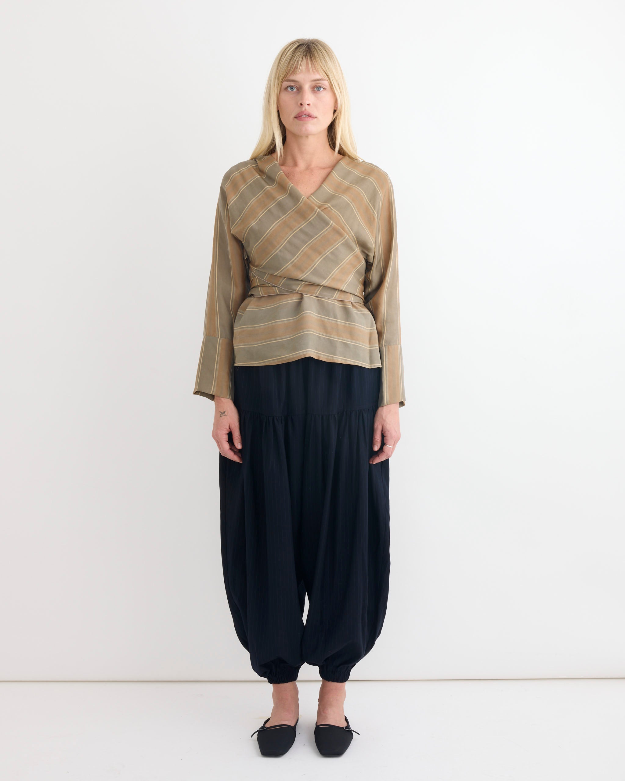 A woman with straight blonde hair and a neutral expression wears the Chelsea Mak Fiona Top in Tie Stripe, paired with loose black harem pants gathered at the ankles and black pointed shoes, standing against a plain white wall.