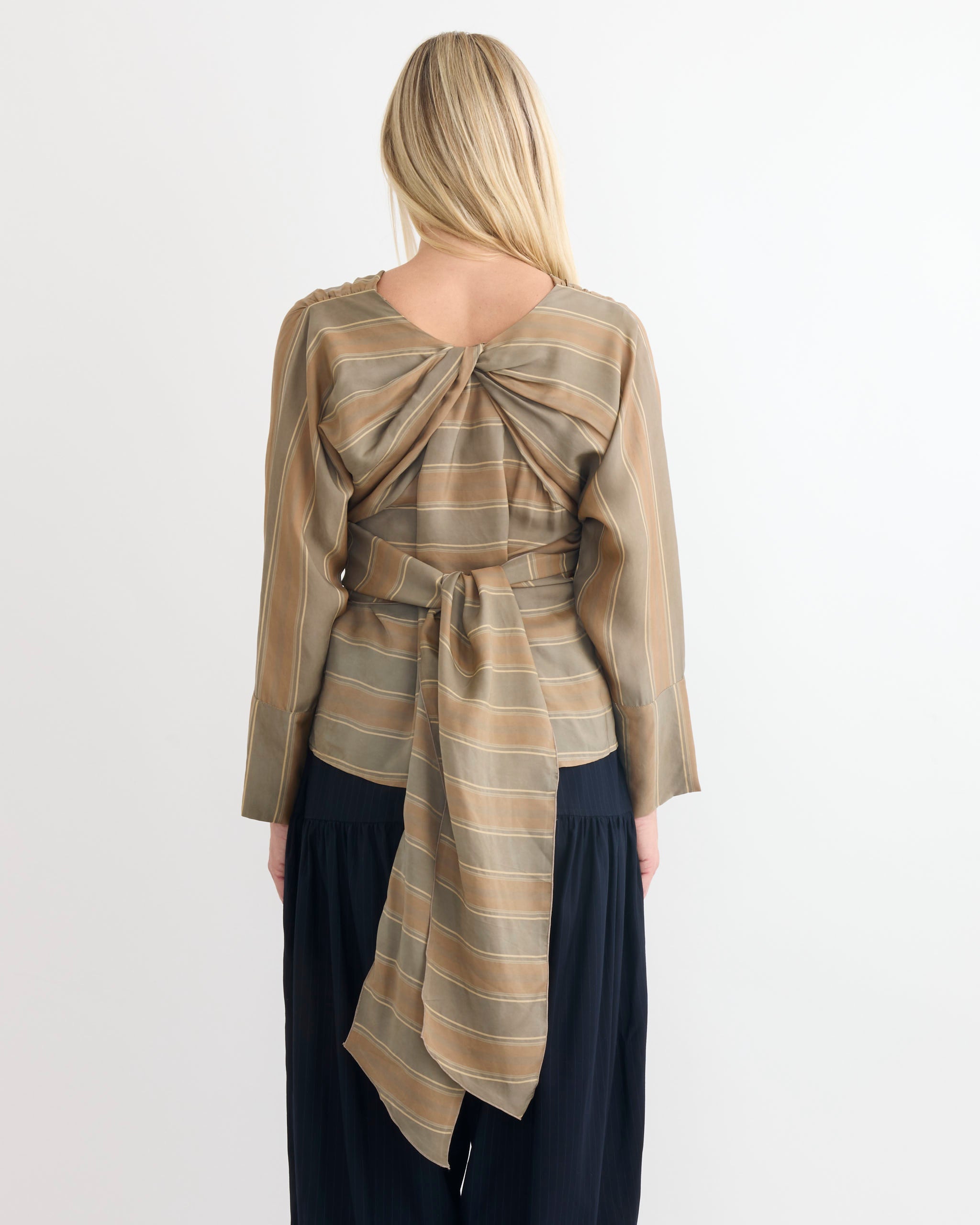 A woman with long blonde hair is seen from the back, wearing the Chelsea Mak Fiona Top in Tie Stripe, featuring a wrap style and tie detail, paired with dark pleated pants.