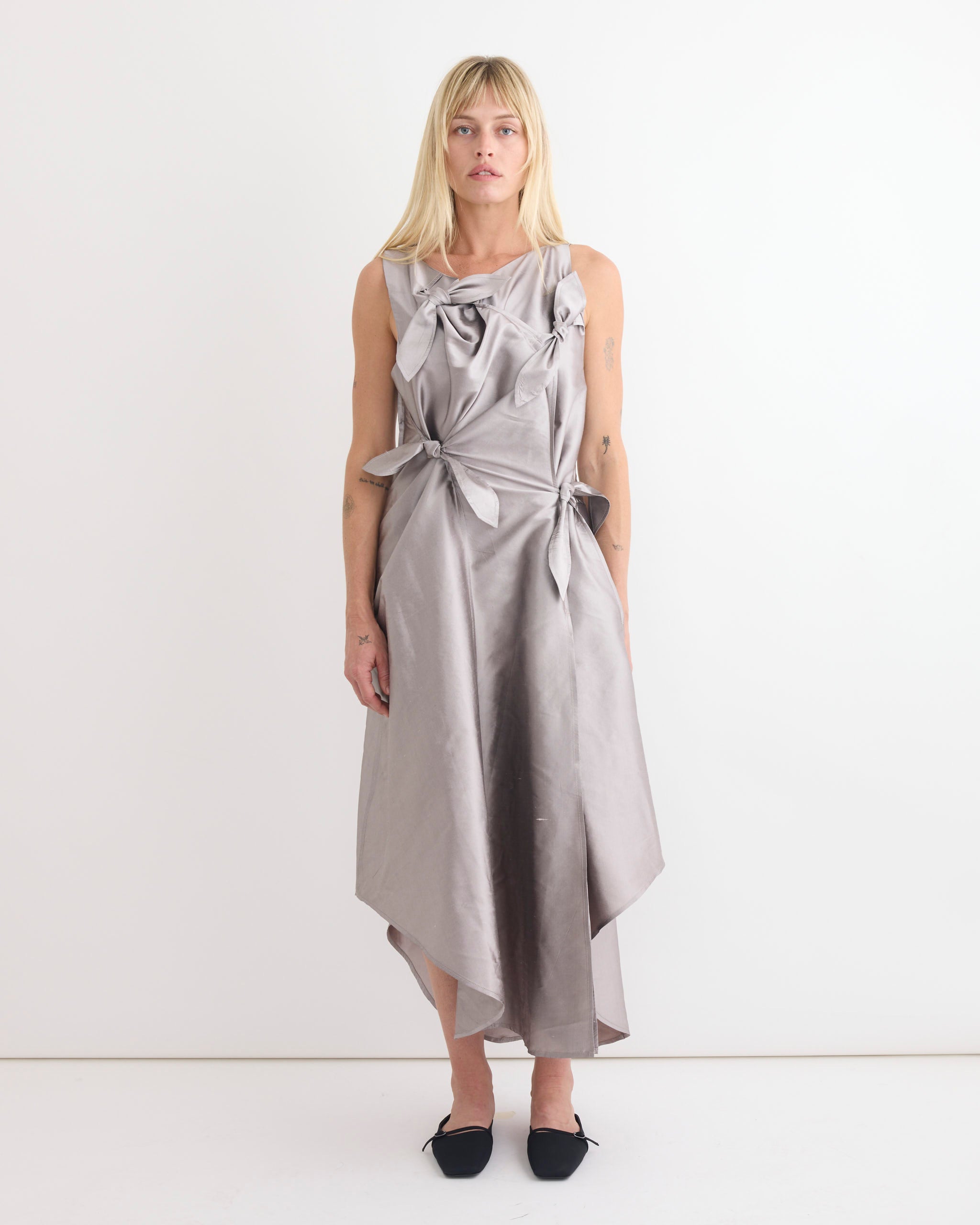A woman with straight blonde hair stands against a white background in the Chelsea Mak Tabea Dress in Lavender Taffeta, a sleeveless, asymmetrical tie-front dress, styled with black pointed-toe flats.