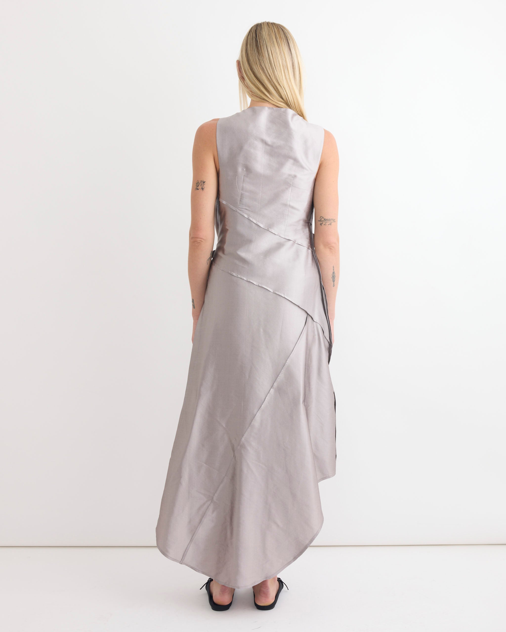 A person with long blonde hair, in the sleeveless, lavender Chelsea Mak Tabea Dress in taffeta and black heels, stands facing a white wall, highlighting the asymmetrical back of this outfit.