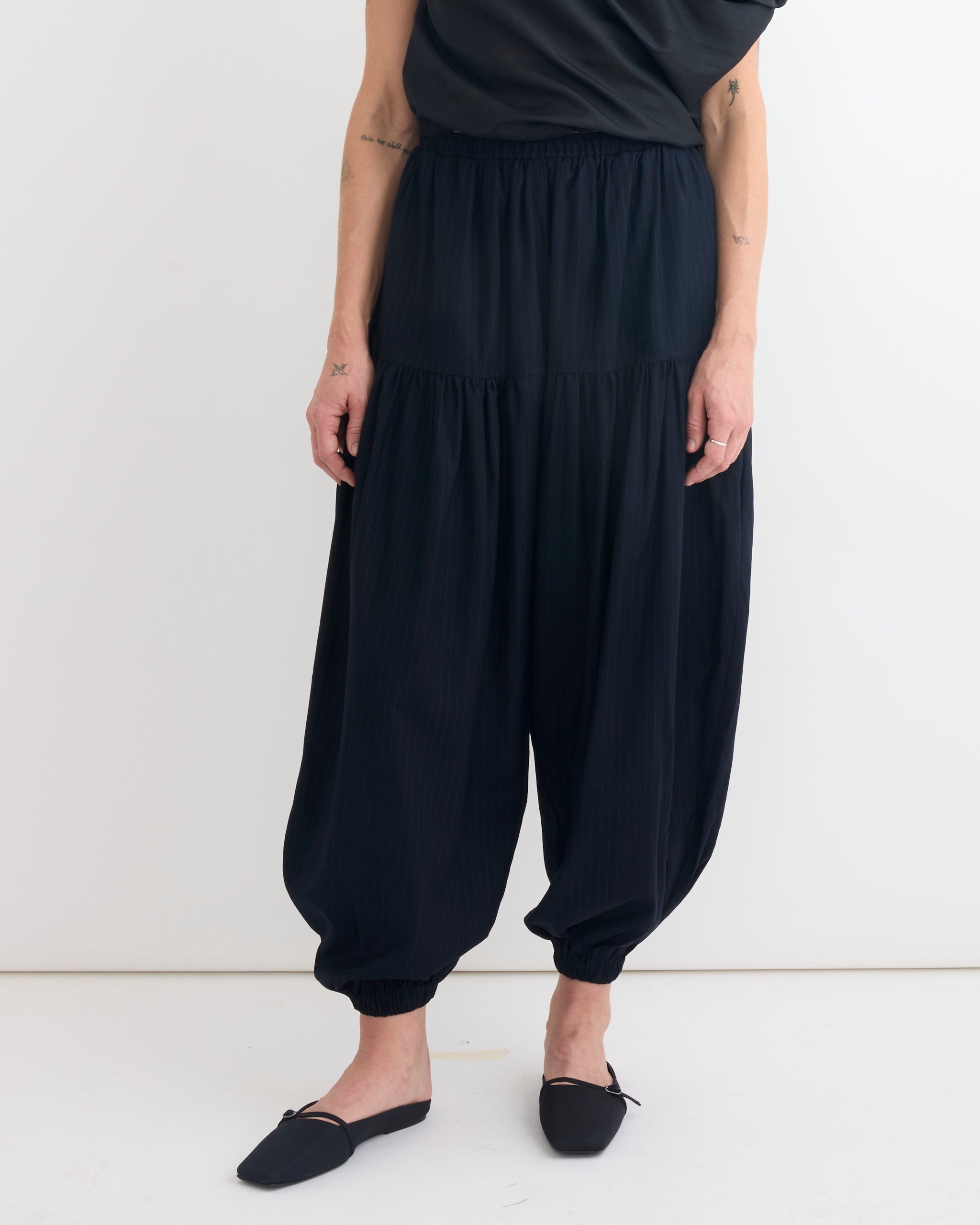 A person wears Chelsea Mak's Rain Jogger in Pinstripe—loose, high-waisted pants with gathered ankles—paired with a black top and flat shoes, standing against a plain white background with arms relaxed at their sides.