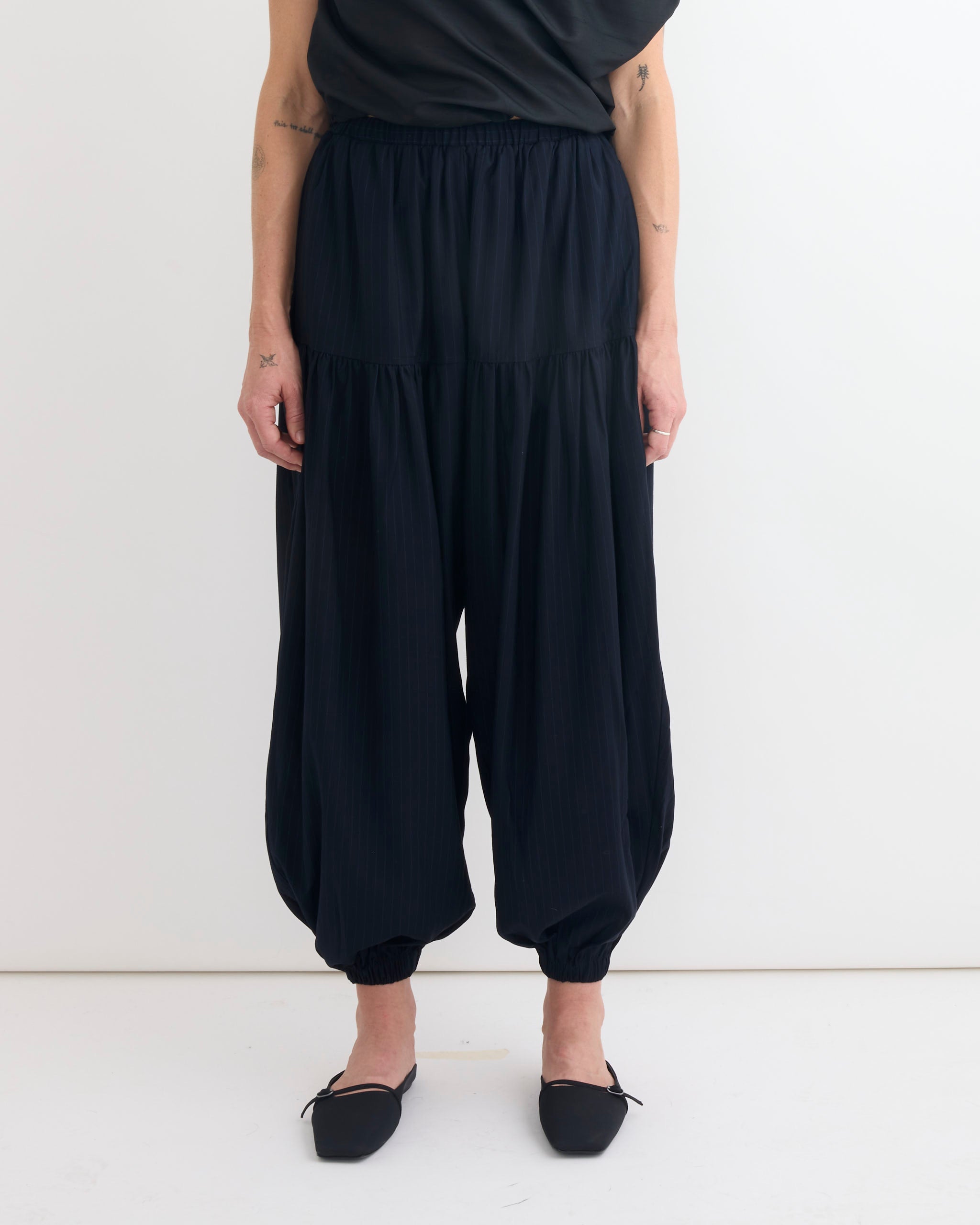 Only the lower half of a person wearing Chelsea Mak’s Rain Jogger in Pinstripe is visible. The loose, balloon-style jogger pants with elastic cuffs are paired with black flat shoes and a black top, standing against a plain white background.