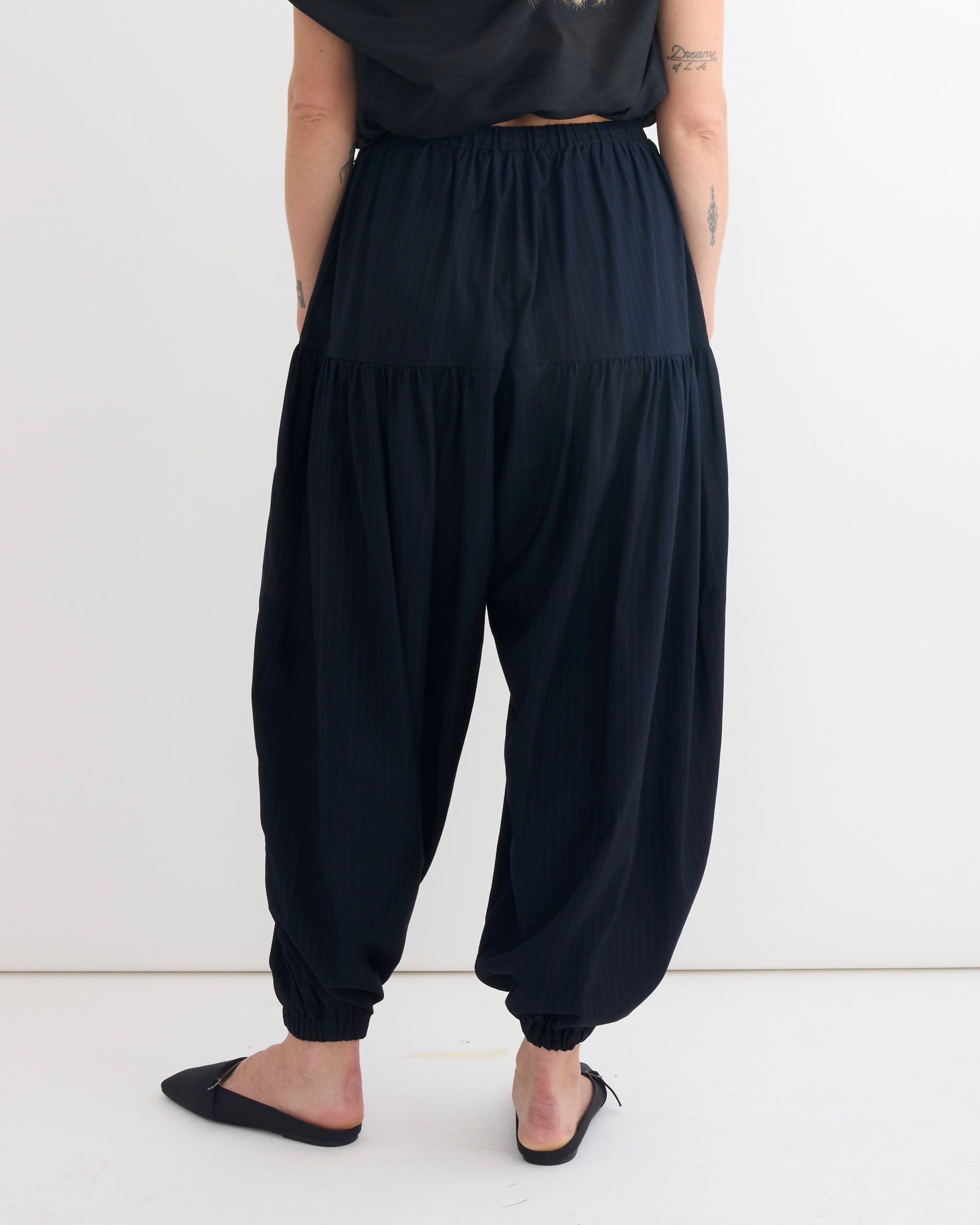 A person stands barefoot against a white backdrop, wearing Chelsea Mak's Rain Jogger in Pinstripe with gathered cuffs and a black top. Their partially visible arms feature several small tattoos.